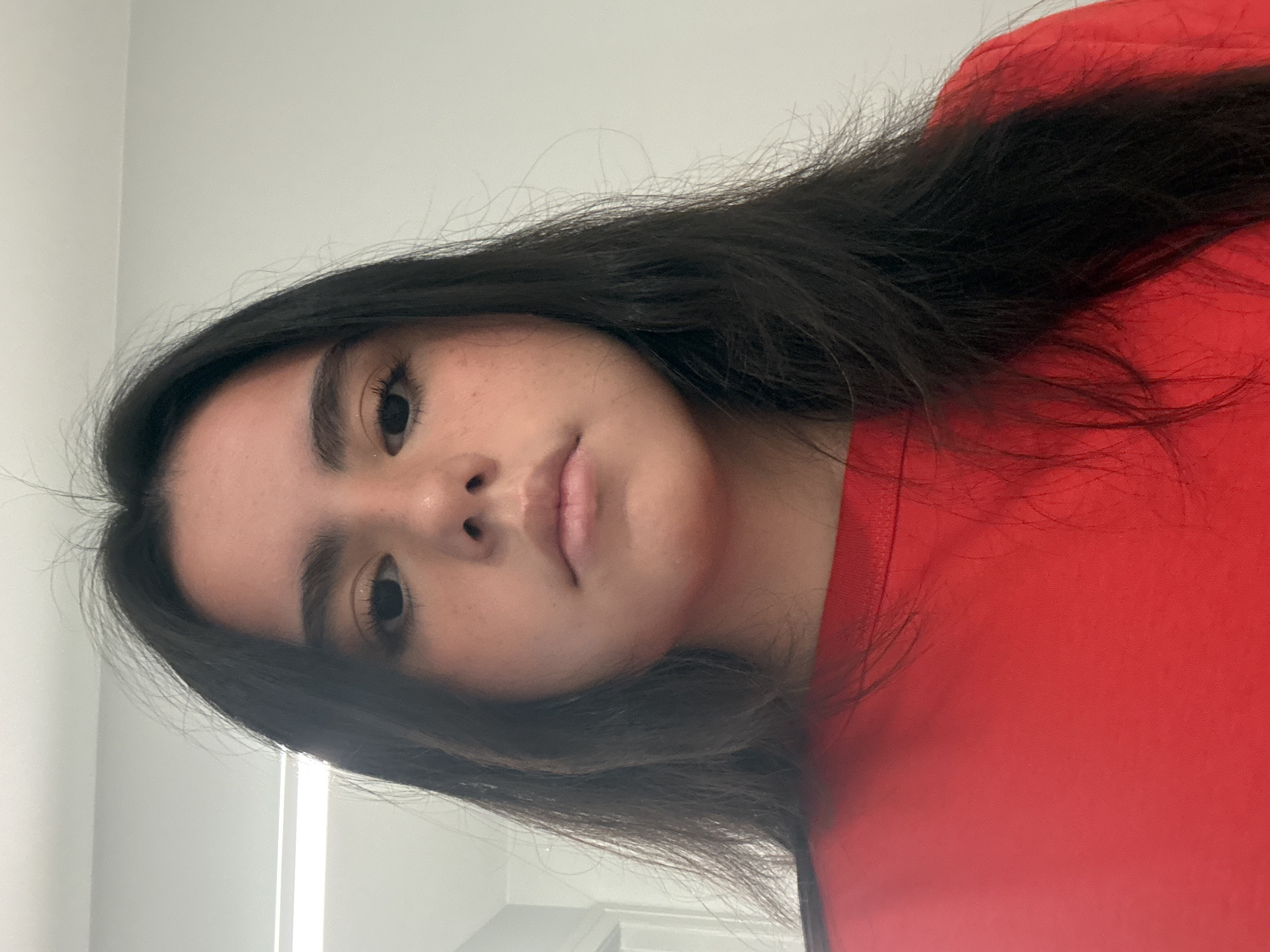 A person with dark hair and a red shirt is facing the camera indoors. The background is a plain white wall. The person's face is visible, showing their eyes, nose, and mouth. The location is not possible to discern from the given image.