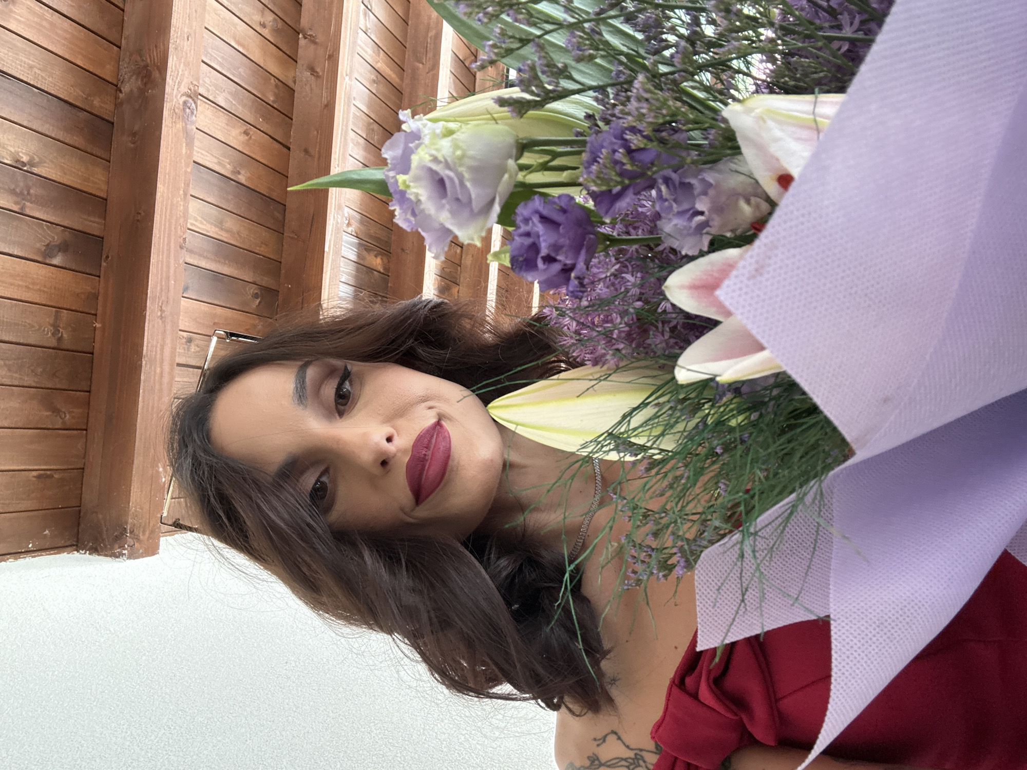 A woman with dark hair and red lipstick is holding a bouquet of lilies and purple flowers. The woman is facing forward, slightly tilted to the left. The background shows a wall with wooden planks and a white surface. The location is possibly Ribaritsa, Bulgaria, based on the user's prompt.