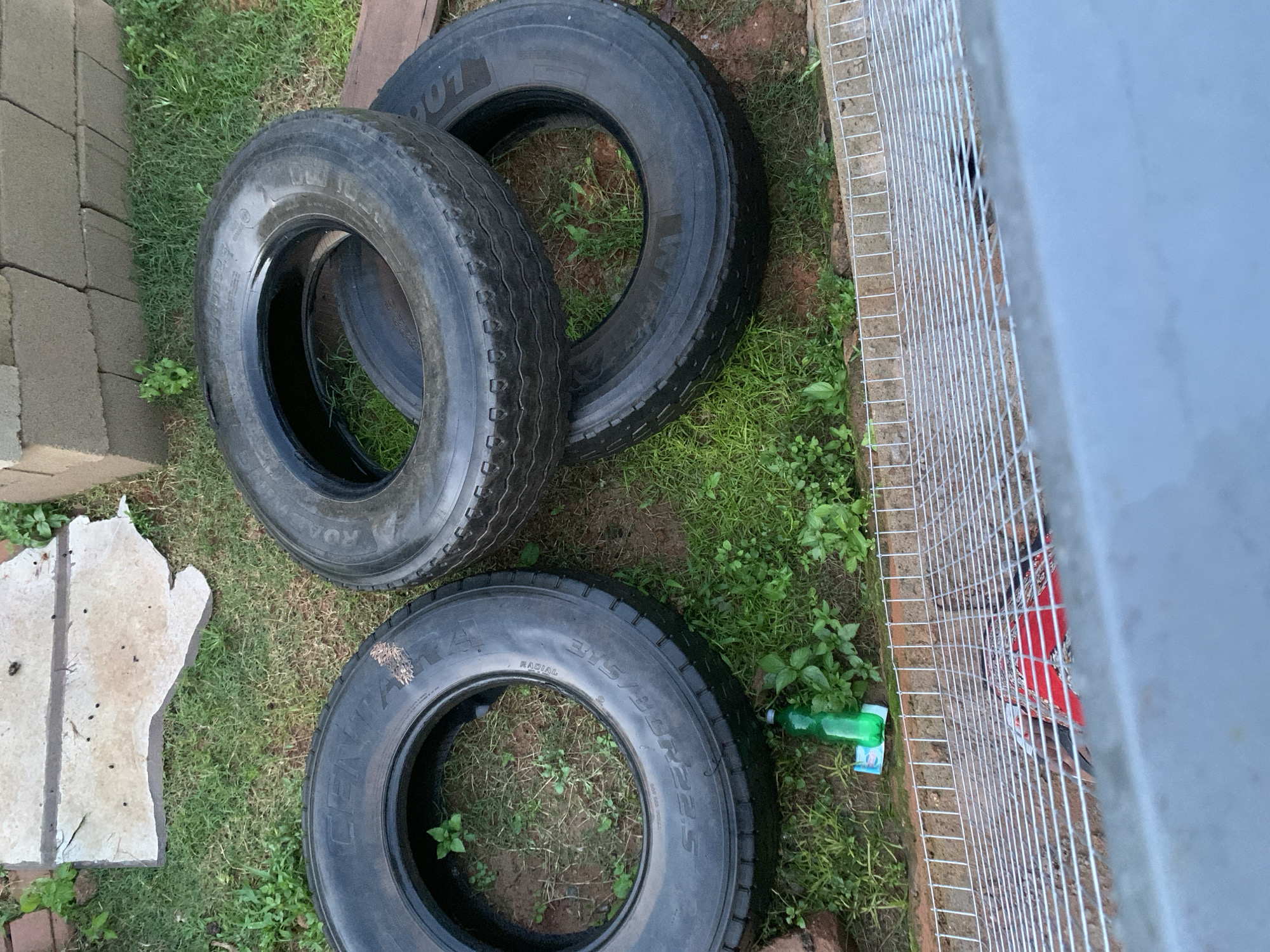 Three used tires are stacked on grass. The top tire is larger than the tire below it, and the third tire is placed next to the stack. In the background, a wall made of concrete blocks is visible. A green plastic bottle and a small white packet are on the grass near the tires. In the foreground, a wire mesh fence is present, with a glimpse of a red object underneath.