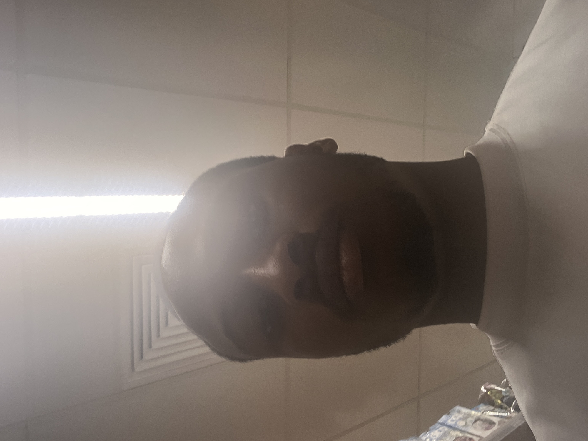 A close-up, low-angle shot of a man indoors. The man has dark skin, a shaved head, and a trimmed beard. He is wearing a white shirt. The background is composed of white ceiling tiles and a bright fluorescent light. The location cannot be determined from the image.