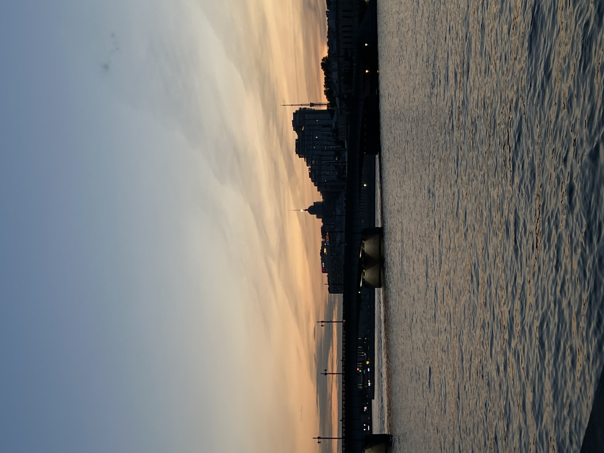 This is a horizontal, eye-level, outdoor, full-shot photograph of a bridge over a body of water at dusk. The sky is a gradient of light blue at the top to pale orange and yellow near the horizon, with thin, wispy clouds. A silhouetted cityscape with various buildings and a tall tower is visible on the far bank, beyond the bridge. The bridge itself is dark and appears to be made of concrete or stone. Lights are visible under the bridge and on the distant buildings, casting reflections on the water. The water is rippled and reflects the colors of the sky and the lights. The overall mood is peaceful and serene. The location is Unknown, Russia.