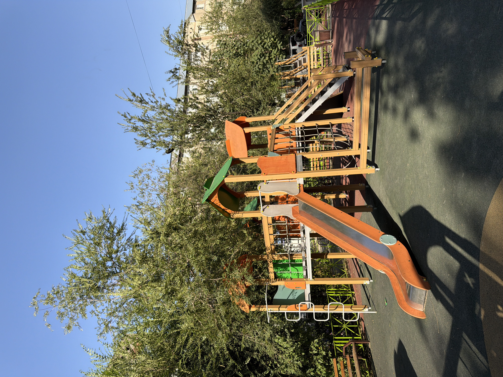 A brightly lit, outdoor children's playground is depicted on a clear, sunny day. The playground features a large wooden structure with orange and green accents, including two slides, stairs, and climbing nets. The ground surface appears to be a mix of dark green and red rubberized material, common in playgrounds for safety. Lush green trees surround the playground, with their branches partially obscuring the clear blue sky. In the background, a multi-story apartment building is visible, suggesting an urban or suburban setting. A wooden bench is placed to the left of the playground structure. The scene is free of people or any activity. This playground is located in the city of Shymkent, Kazakhstan.