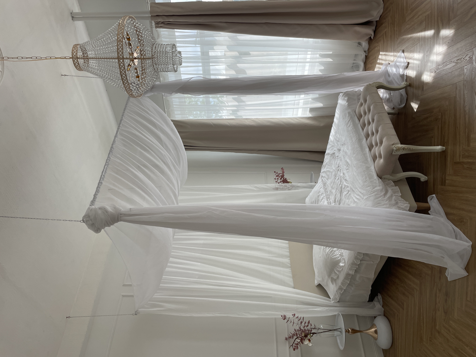 A bright, naturally lit interior space contains a four-poster bed draped with white, semi-transparent fabric. A crystal chandelier hangs above. A tufted cream-colored chaise lounge sits at the foot of the bed on a wood-paneled floor. Sheer curtains cover the window, allowing natural light to filter in. Two small tables, each holding a vase with dried flowers, flank the bed.
The location is Kazan, Russia.