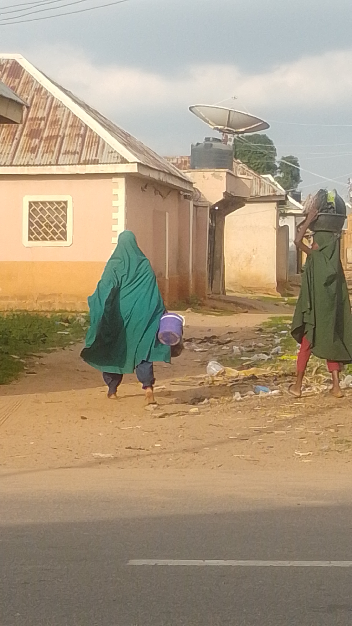 The image depicts a scene in Tempure, Nigeria, featuring two individuals walking on a dirt path beside a road. The person in the foreground, facing away from the camera, is wearing a large, flowing emerald green garment that covers their head and body, with blue trousers and sandals visible. They are carrying a purple bucket in their right hand. Further down the path, a second individual, also facing away, is wearing a similar green garment and red trousers. They are carrying a dark, cylindrical object balanced on their head.
The environment appears to be a semi-urban or rural area, with simple, single-story buildings in the background. One building has a corrugated metal roof, and another has a satellite dish on its roof. The path is unpaved and appears somewhat littered with plastic debris. A paved road runs in the foreground of the image. The sky is overcast with clouds.
