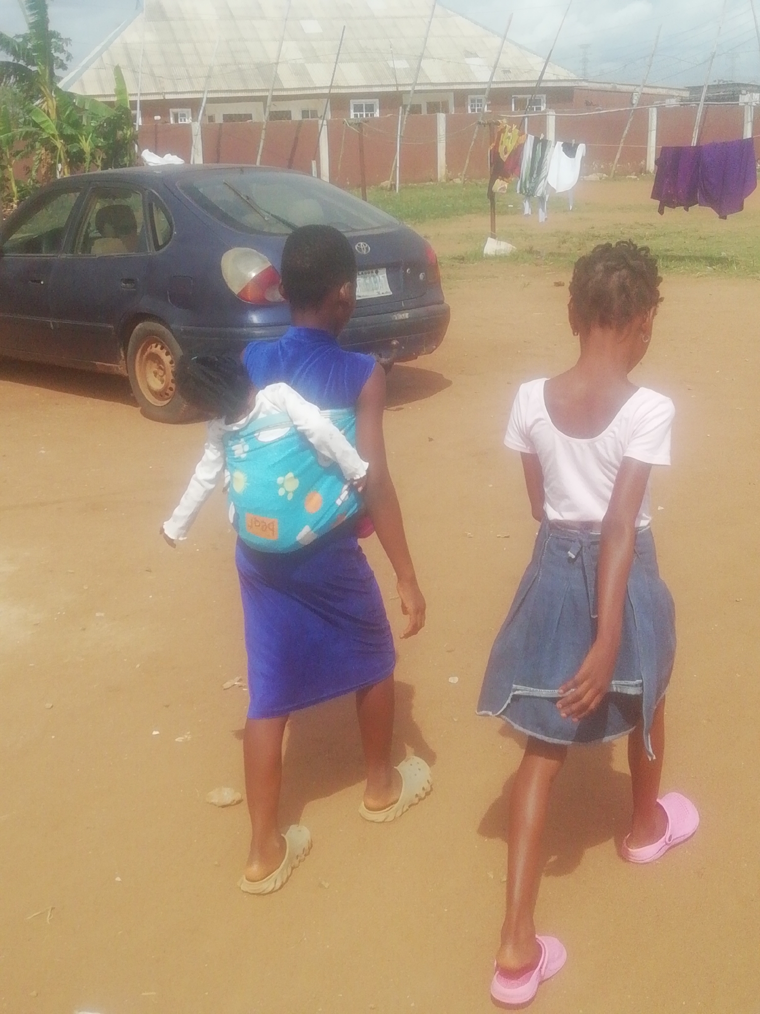 Two young girls walk on a dirt road in Ikorodu, Nigeria. The girl on the left wears a blue dress and carries a child in a blue and green carrier on her back. The child wears a long-sleeved white shirt. The girl on the right wears a white shirt and denim skirt, and pink shoes. To the left is a navy-blue car. In the background, there is a building with a white roof. Laundry hangs on a clothesline. The ground is dirt.