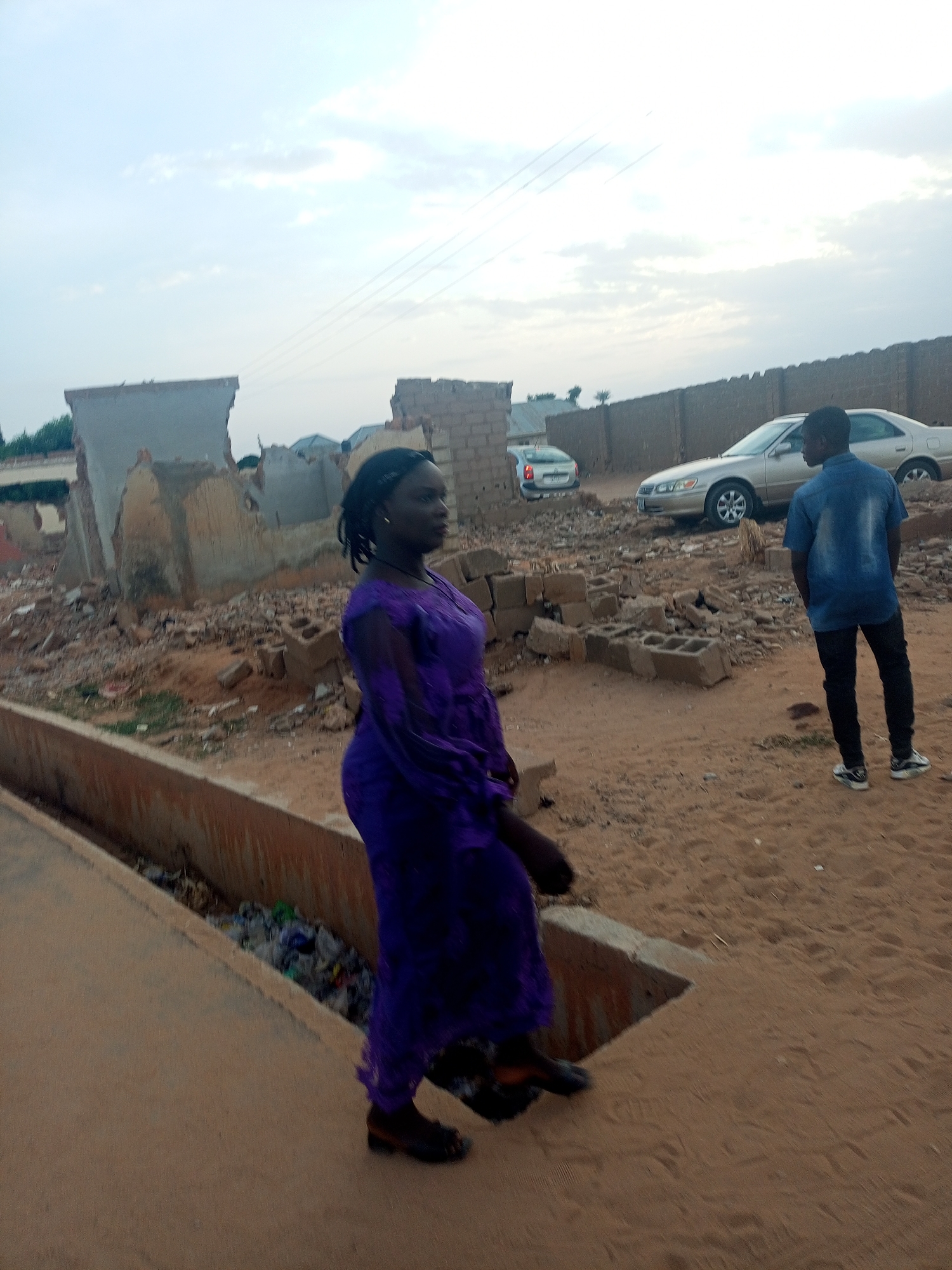 A medium shot depicts a woman in a full-length purple dress walking on a concrete path in a sandy area, with a man standing in the background. The background includes dilapidated structures, a wall, and two cars. The sky is overcast.