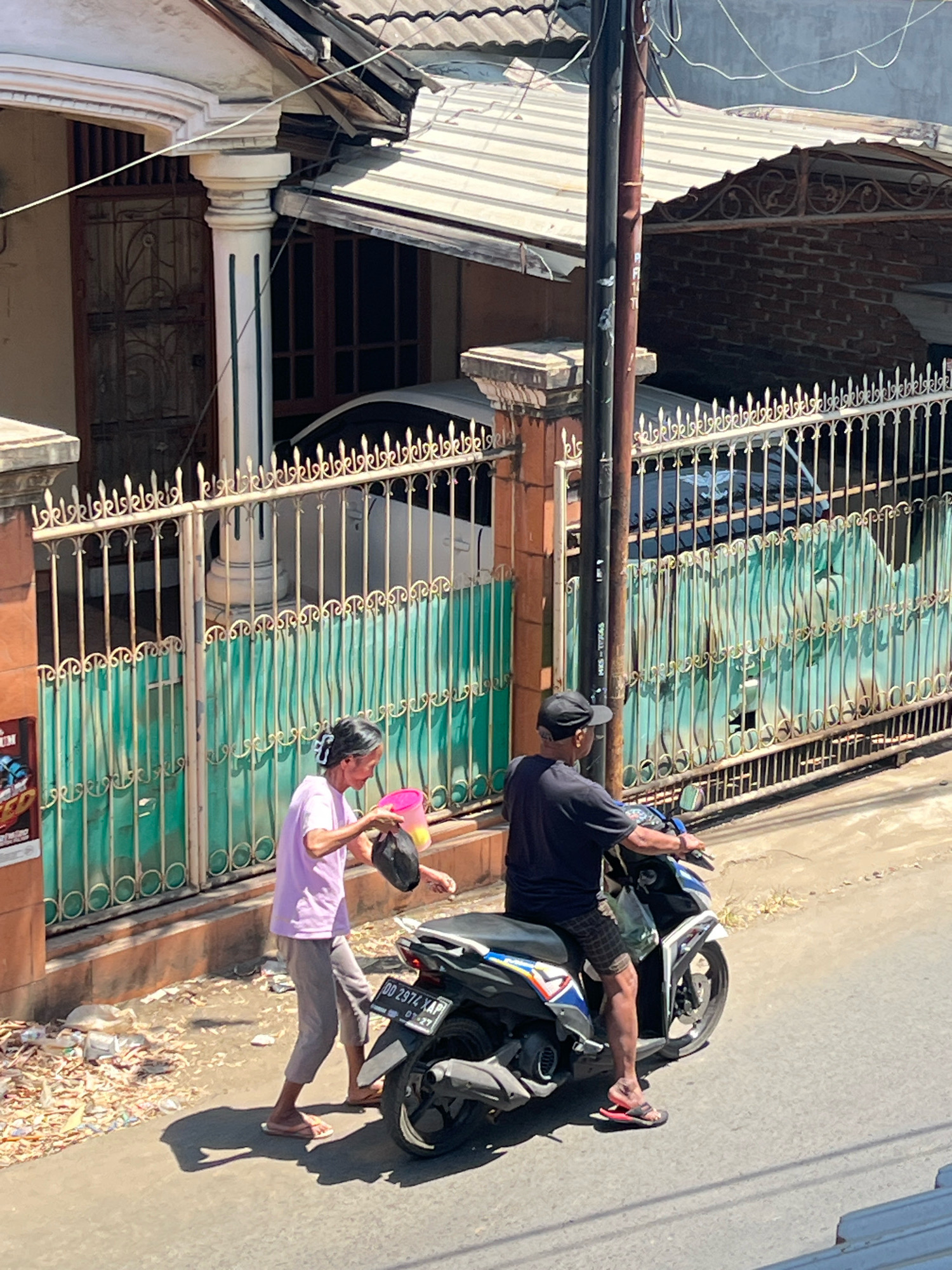 In Makassar, Indonesia, on a sunny day, a woman in a purple shirt and grey pants walks on a paved road. She carries a pink bucket in her right hand and a black bag in her left. Beside her, a man wearing a black t-shirt and a black cap rides a motorcycle. He is looking forward, and the motorcycle is partially obscuring the woman from view. Behind them, a white and green fence encloses a property with a white car parked inside. The scene is set in front of a house with a tiled roof and a decorative facade. The road is lined with scattered debris, suggesting a somewhat unkempt environment.
