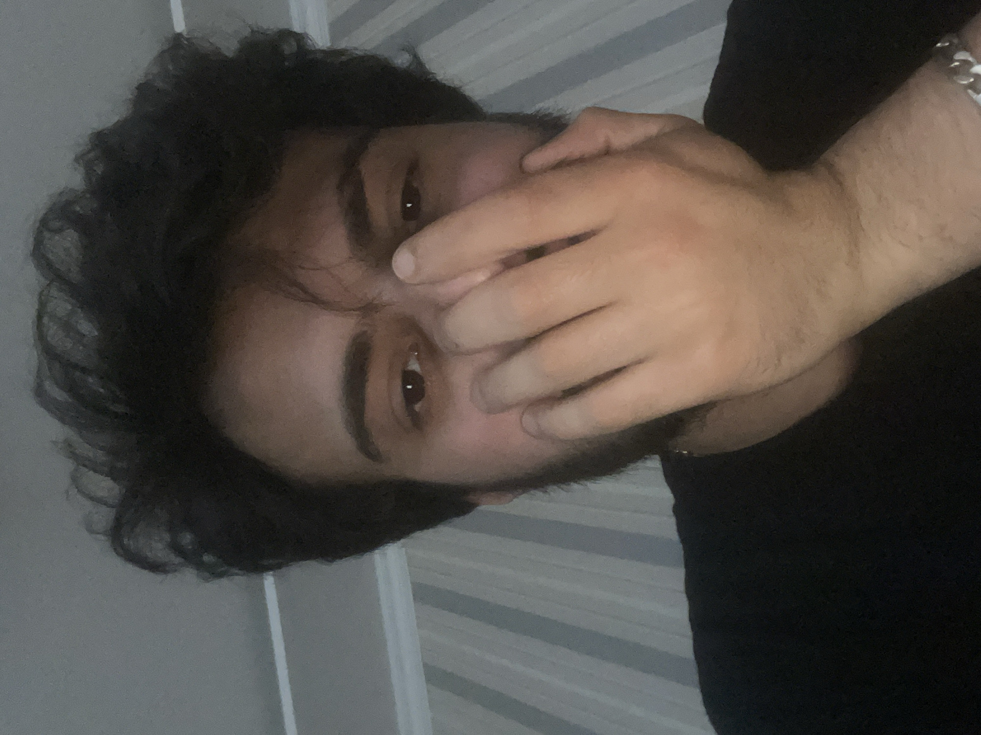 The image is a close-up, indoor selfie of a person's face and chest. The subject has dark, curly hair and is wearing a black t-shirt. Their right hand is raised to cover their nose and mouth, obscuring the lower half of their face. Their eyes are visible, and they appear to be looking directly at the camera. The background consists of striped wallpaper and a ceiling. There is no discernible information in the image to indicate the location is Tbilisi, Georgia.