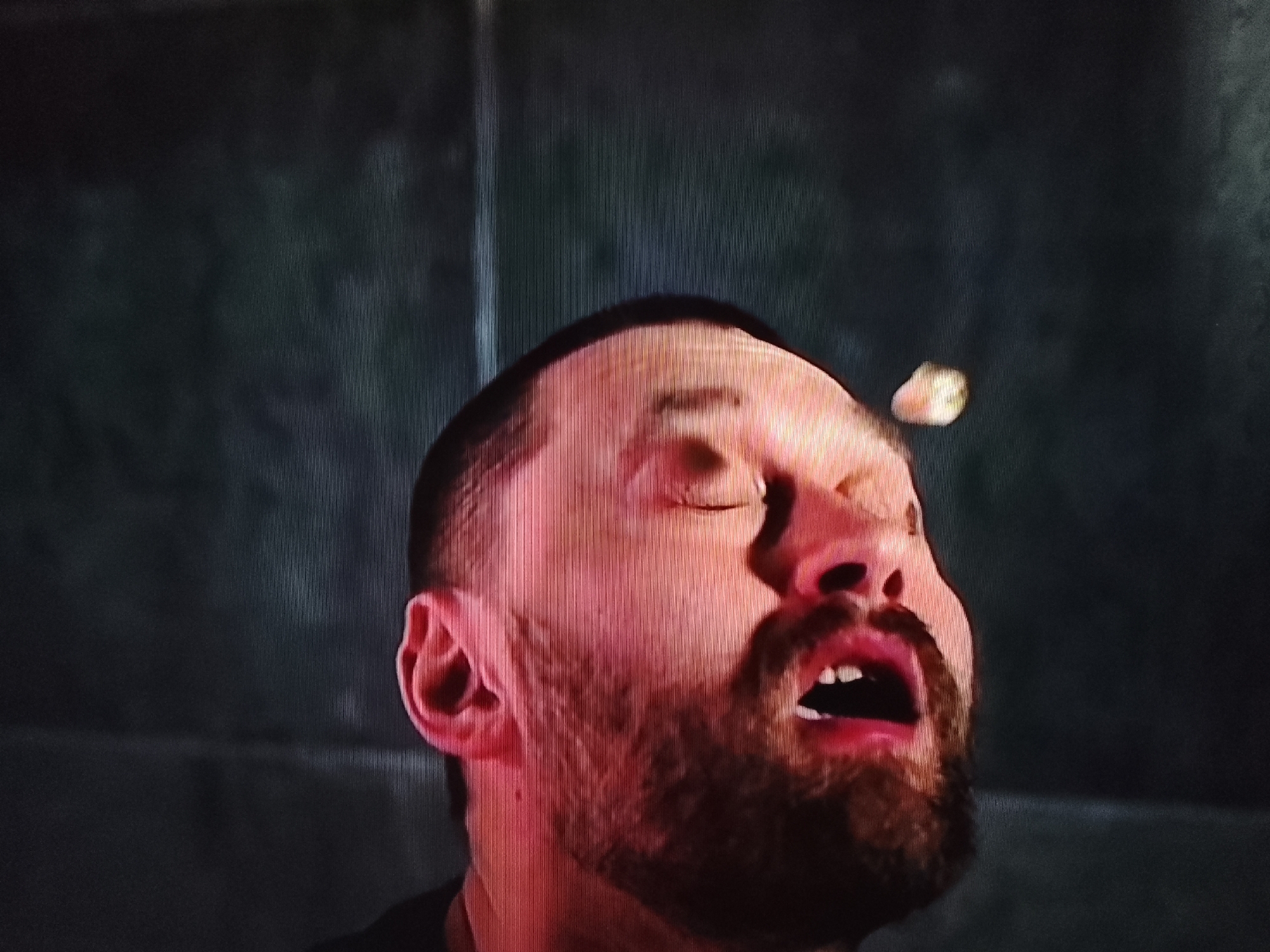 The image shows a close-up, slightly blurred shot of a man's face with his eyes closed and mouth open. A small, light-colored object is in mid-air near his forehead. The man has short dark hair, a beard, and is illuminated by red lighting. The background is dark and textured. There is no discernible location in the city of Klichaw, Belarus.