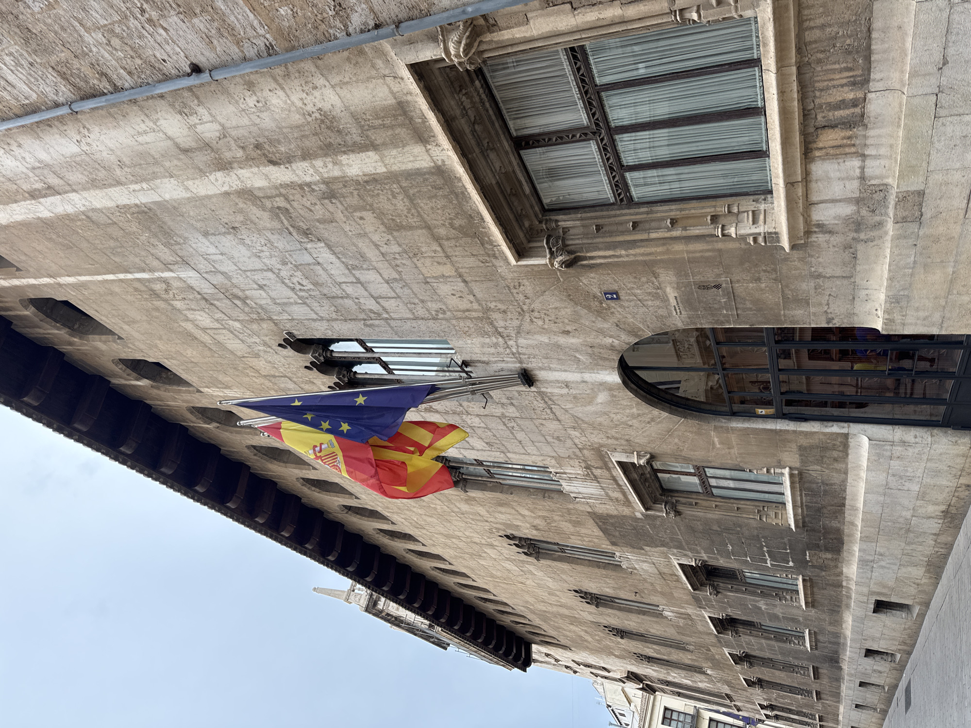 The image shows the façade of a historic stone building in Valencia, Spain. Two flags, the Spanish flag and the European Union flag, are prominently displayed, hanging from a pole mounted on the building. The building features a series of arched windows and decorative stone carvings. A large arched doorway with glass doors is visible on the right side of the image. The lower part of the building is made of large stone blocks, and a sidewalk runs along the front. The sky is overcast. The building's architecture suggests it is a significant public or administrative building. The signage on the wall below the European flag includes the number "2", the words "Justícia Pal de la Generalitat", and a coat of arms, identifying the location as the Palace of Justice of the Generalitat of Valencia.
