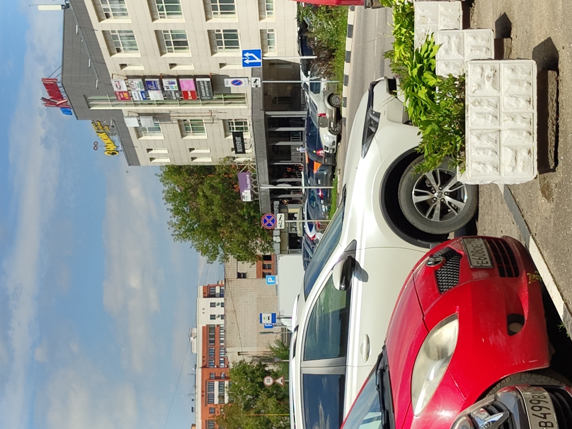 The image captures a sunny day in Zhukovsky, Russia, focusing on a street scene. In the foreground, a red car and a white SUV are parked along a sidewalk. The white SUV's right rear tire is visible, along with a planter box filled with green foliage. In the background, a multi-story building with a gray facade and many windows dominates the skyline. Several signs are affixed to the building, including a red sign with Cyrillic text at the top. Trees and other buildings are also visible further in the distance. Street signs indicating parking and traffic regulations are present. The sky is blue with scattered white clouds.