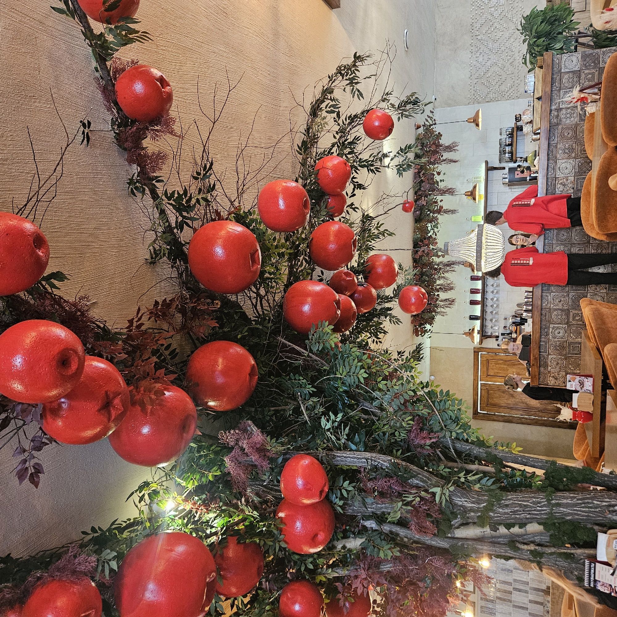 The image shows the interior of a cafe decorated with artificial trees and large red fruit ornaments hanging from the ceiling. There is a bar area with several people, including two employees wearing red shirts and one employee in black. The cafe has orange seating and is illuminated by pendant lights and a chandelier. The restaurant is located in Chelyabinsk, Russia.
