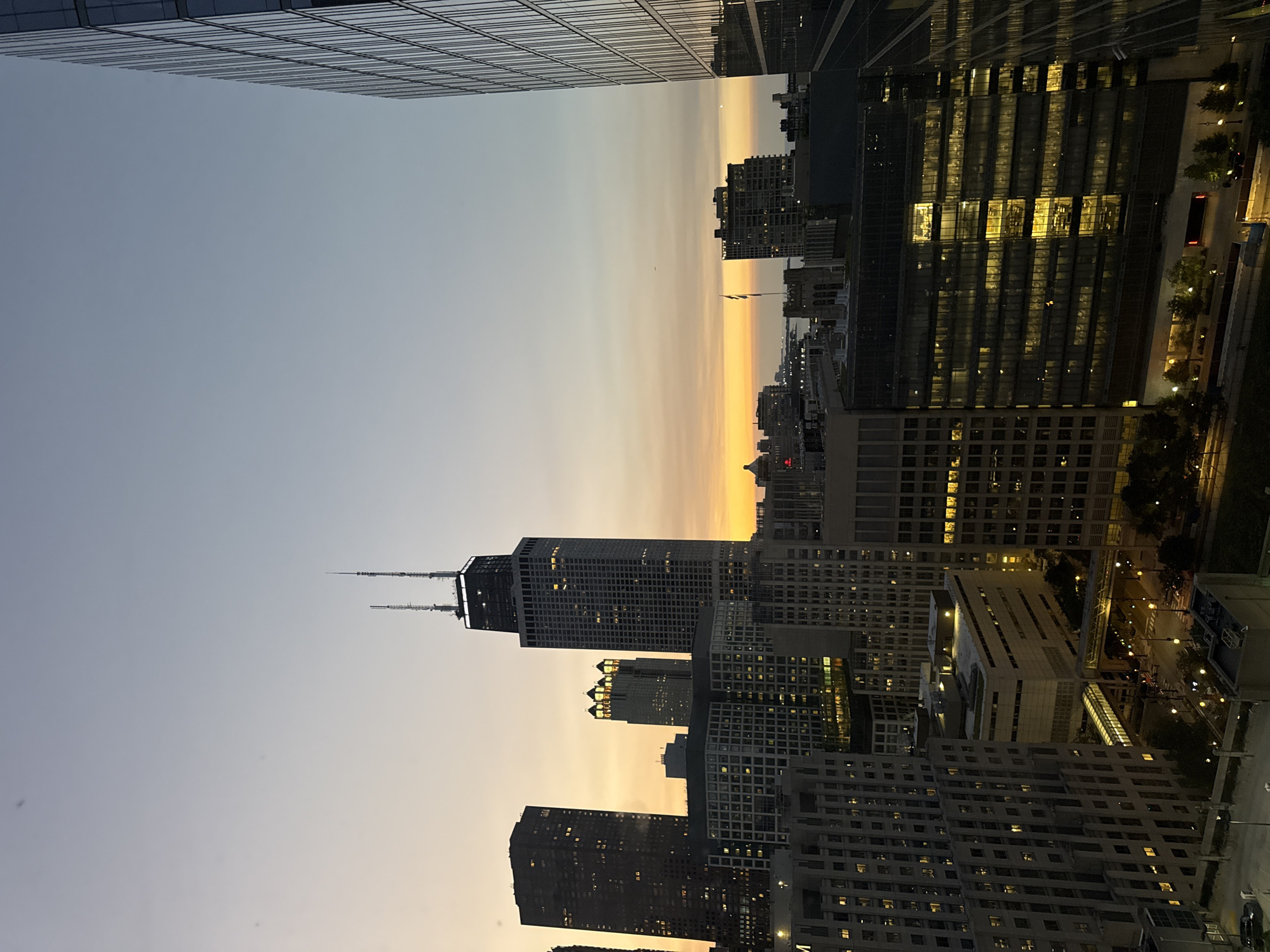 The image shows a cityscape of Chicago at dusk, featuring several skyscrapers. In the center, the John Hancock Center, a prominent Chicago landmark, is visible with its distinctive cross-braced structure and twin antennas. The sky displays a gradient of colors from light blue at the top to orange and yellow near the horizon, indicating the setting sun. Various other buildings of different heights and architectural styles surround the John Hancock Center. The foreground and midground show a dense urban environment with buildings, some illuminated from within, suggesting office spaces or residences. The overall scene captures the vastness and verticality of a major metropolitan area as daylight fades.