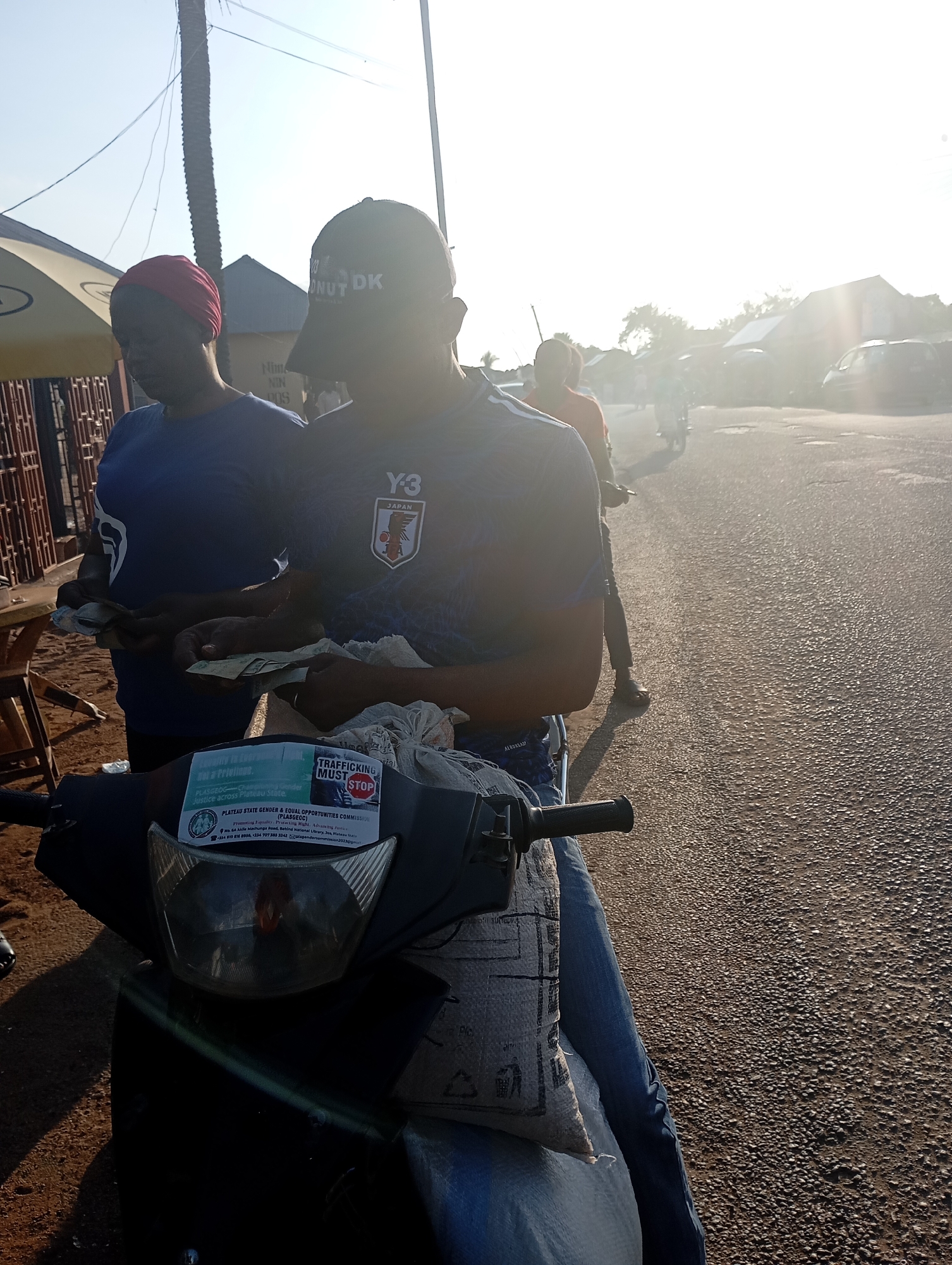 Two individuals, one wearing a red headscarf and a blue t-shirt, the other wearing a black baseball cap and a dark blue t-shirt with a Japanese anime-style logo, are interacting in front of a parked motorcycle. The person in the red headscarf appears to be handing or receiving money from the person in the baseball cap. The motorcycle has a sticker on the front that reads "TRAFFICKING MUST STOP" and contains information about the "PLATEAU STATE GENDER & EQUAL OPPORTUNITIES COMMISSION (PLASGEOC)". The scene is outdoors, with a paved road and buildings in the background. The lighting suggests it is daytime, possibly morning or late afternoon, with the sun creating a bright glare in the upper right portion of the image. The environment appears to be a street or roadside area within the city of Shendam, Nigeria.
