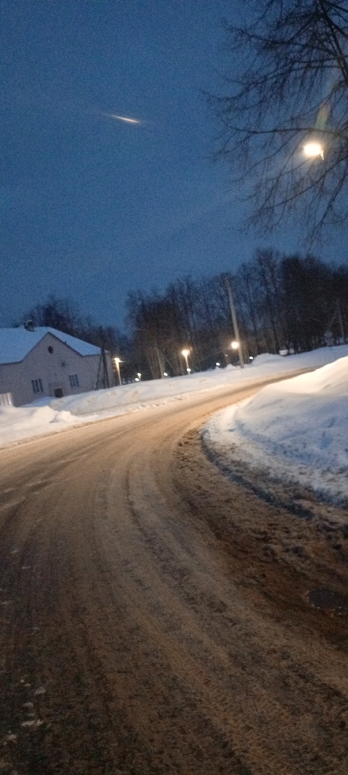 The image depicts a winter street scene at dusk or night in Красный Маяк, Russia. A curved, partially snow-covered asphalt road, displaying visible tire tracks and areas of dirt/slush, dominates the foreground, winding from the lower center towards the upper right. Snowdrifts line both sides of the road. To the left, a two-story, light-colored building with a dark gabled roof and multiple windows is visible, partially obscured by snow. A line of bare, leafless deciduous trees forms a dark silhouette across the midground and background, behind which additional streetlights are visible. Multiple active streetlights, emitting a warm yellow-orange glow, illuminate sections of the road and trees. The sky is a dark, deep blue, and a distinct bright, elongated streak of light is present in the upper left quadrant. No people or vehicles are visible.
