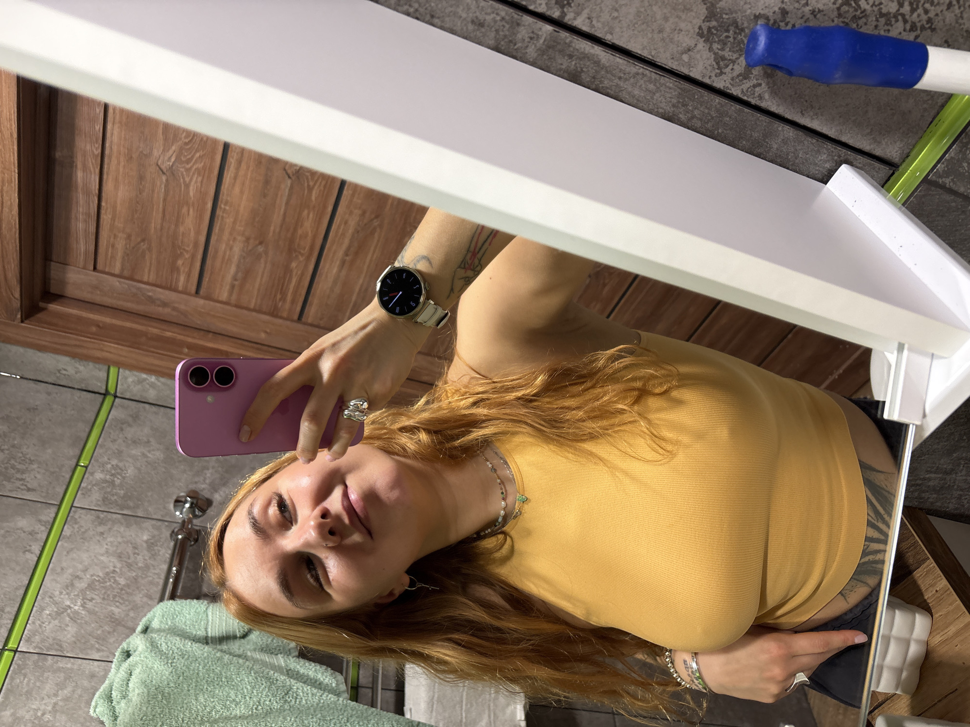 The image shows a person with fair skin and long, wavy blonde-red hair taking a selfie in a bathroom mirror. The person is wearing a fitted yellow top, a watch on their left wrist, multiple rings on their fingers, and necklaces. Tattoos are visible on both arms. The pink smartphone being used to take the photo has two lenses.
The bathroom features a wooden vanity cabinet, green patterned tiles, a gray tiled floor, a mint green towel, and a chrome faucet. A cleaning tool with a blue handle and green stem is visible in the background. The exact location in Minsk, Belarus is not discernible from the image.
