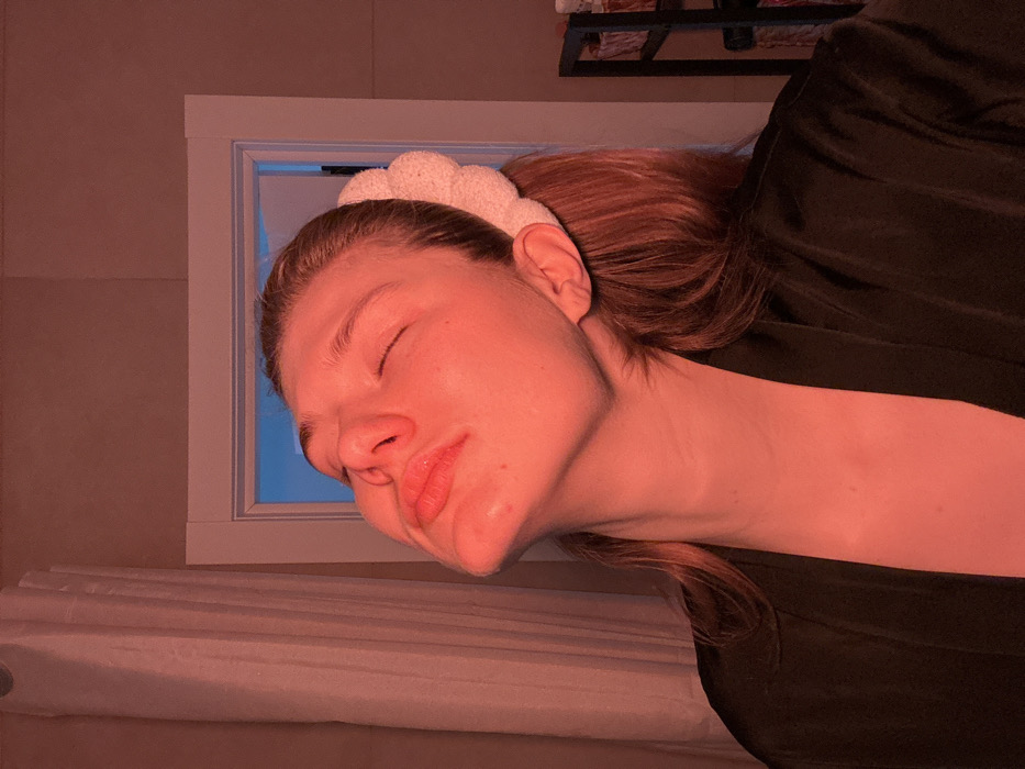 The image depicts a female individual indoors in Nikol'skii, Russia. The subject, with light-colored hair, is shown from the chest up, with her head tilted sharply backward, eyes closed, and chin elevated. Her lips are slightly parted. She is wearing a dark, smooth-textured robe. A light-colored, textured headband secures her hair. The scene is illuminated by a strong reddish-orange light source, casting a deep red hue across the subject's face, neck, and the immediate surroundings. To the left of the subject, a vertical light-colored curtain is present. Behind the subject's right shoulder, a white doorframe is visible, through which a light-blue illumination emanates. A dark, multi-tiered shelving unit is partially discernible on the far right.