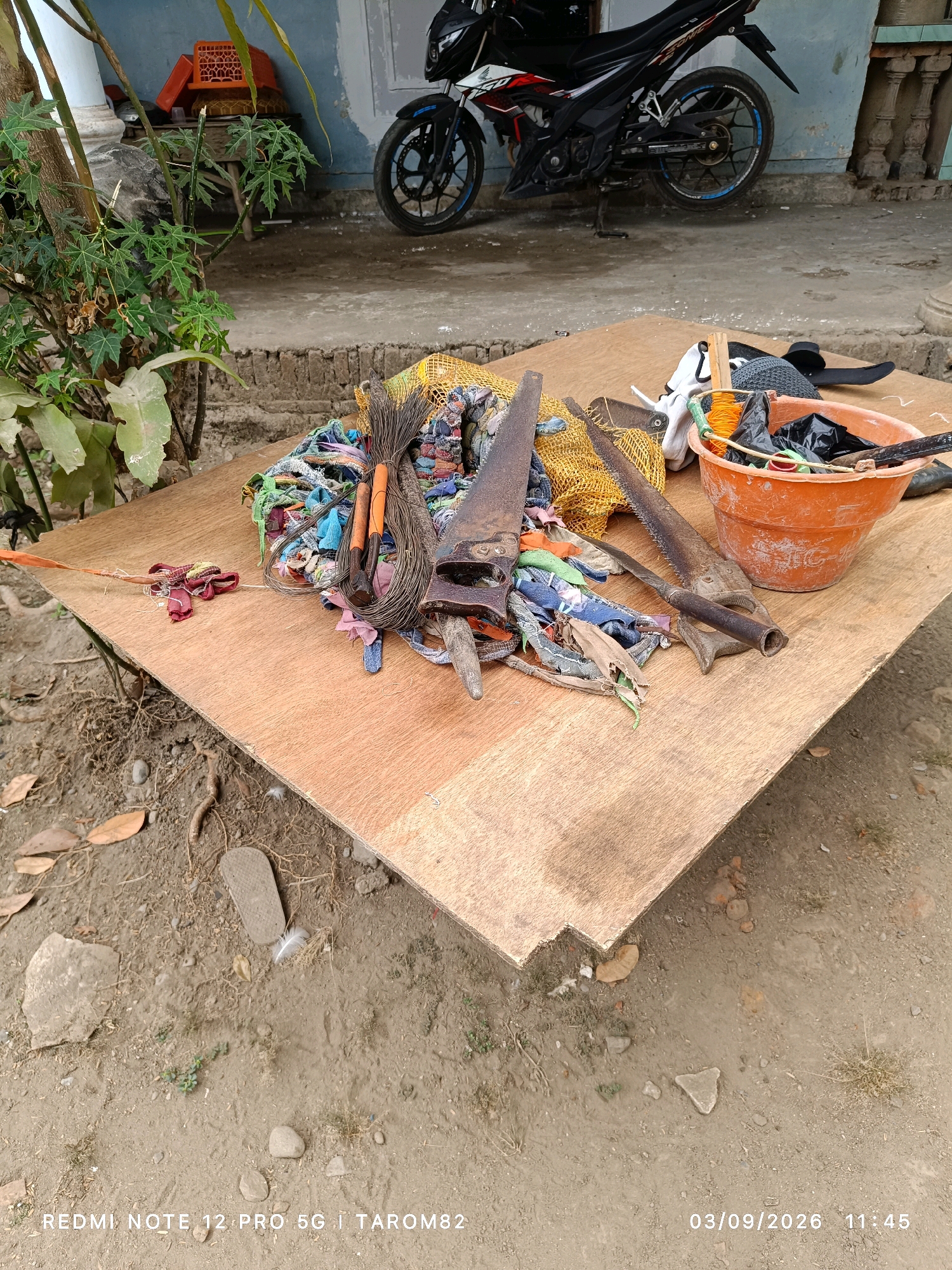 The image depicts an outdoor scene in the city of Rejoagung, Indonesia. In the foreground, a piece of plywood serves as a surface for a collection of objects, including several saws, a bundle of string, and various scraps of fabric. To the right of the plywood, an orange bucket is filled with black plastic bags and some miscellaneous items. In the background, a motorcycle is parked on a concrete surface. A plant with green leaves is visible to the left of the plywood. The ground around the plywood is dirt with some scattered leaves and small stones. The timestamp in the lower right corner reads "03/09/2026 11:45." The text "REDMI NOTE 12 PRO 5G | TAROM82" is present in the lower left corner.