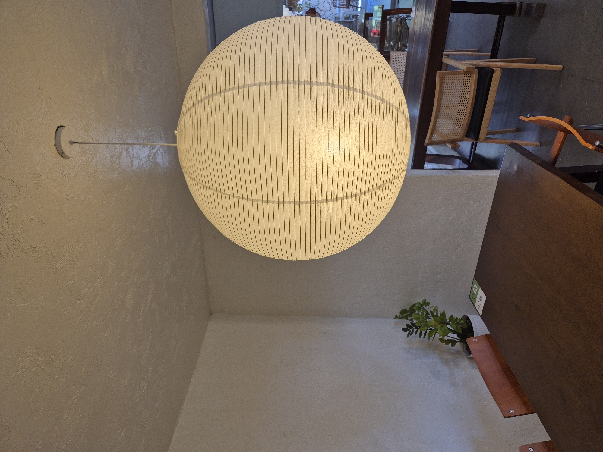 This media file captures an interior scene, likely a cafe or restaurant, in Da Nang, Vietnam. The dominant subject is a large, spherical pendant lamp with a textured, off-white paper shade. The lamp is suspended from a white rod attached to a textured off-white wall. The lamp's surface is characterized by vertical lines, suggesting its construction.
In the background, hints of the establishment's ambiance are visible. Wooden furniture, including chairs with cane weaving and dark upholstery, are arranged around tables. The flooring appears to be a dark, textured material. A wooden partition or wall element is prominent on the right side of the frame. Near this partition, a small potted plant with green foliage is placed on a wooden shelf attached to the wall. There is also a QR code visible on a white background, possibly for menu access or information.
The lighting in the scene is warm and diffused, emanating primarily from the pendant lamp, suggesting it is either evening or the interior is designed for a cozy atmosphere. The textured walls and the choice of furniture contribute to a modern yet natural aesthetic. There are no people visible in the image. The overall mood is one of quietude and contemporary design.