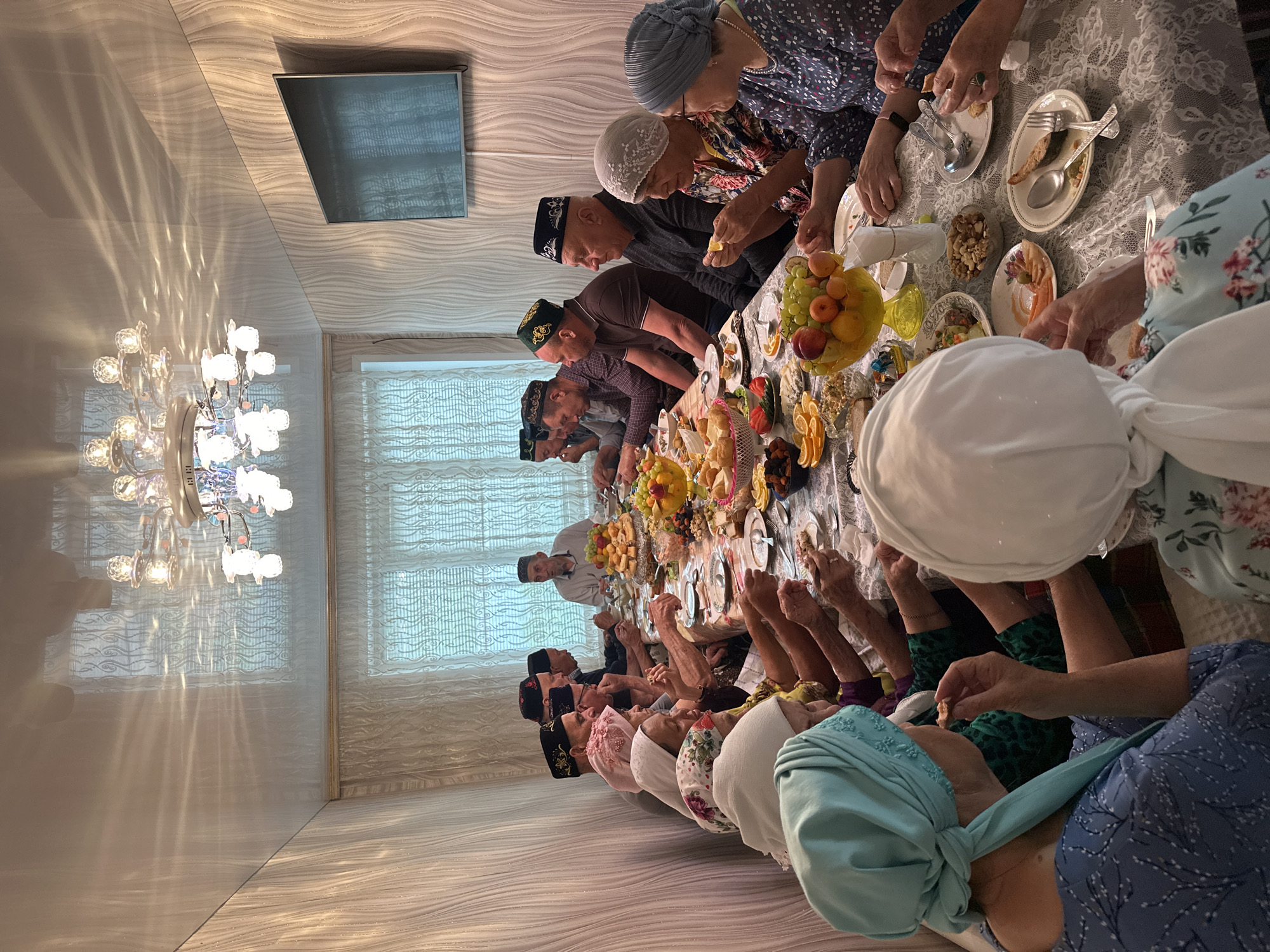 A long table is set for a feast with several people, mostly older men in traditional skullcaps and women in headscarves, seated around it. There is an abundance of food on the table including fruits, nuts, and other dishes. The people are interacting with the food, some eating and others reaching for it. The setting is indoors, in a room that appears to be a dining room with a large chandelier. In the background, there is a window with sheer curtains and a flat-screen TV mounted on the wall. The scene appears to be a gathering or celebration taking place. The location is Коноваловка, Russia.