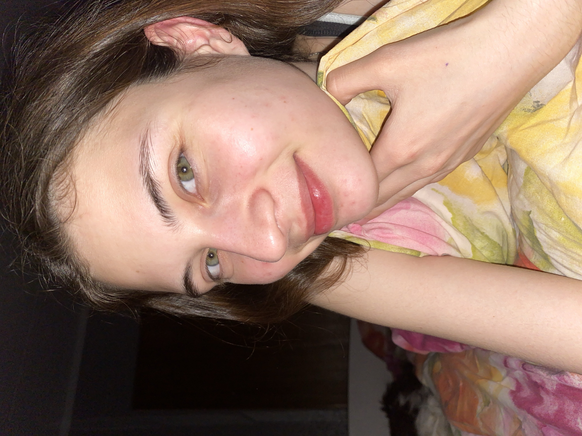 A young woman is pictured in a close-up shot, tilted to the left. Her face is the primary focus, showing her features with a slight smile. She has fair skin, green eyes, and brown hair. Her left hand is near her neck, holding a yellow and pink floral patterned fabric. The background is dark and partially obscured, with only a glimpse of what may be a wall. There is no information available in this image to indicate that the image was taken in Votkinsk, Russia. The location is indeterminable from the available visual data.
