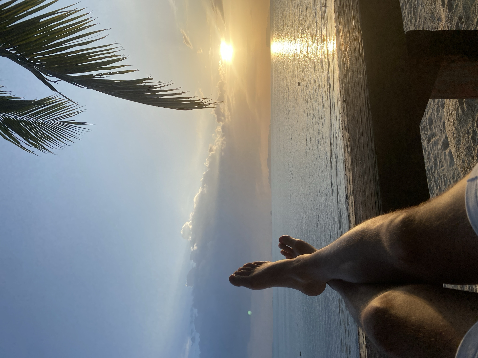 The image depicts a first-person perspective of someone relaxing by the sea at sunset in Koh Samui District, Thailand. The foreground shows the person's bare legs and feet crossed, with their shins and knees illuminated by the warm, golden light of the setting sun. They are seated on what appears to be a wooden bench or railing, with a sandy beach visible below.
The midground is dominated by a tranquil ocean, reflecting the vibrant colors of the sunset, creating a shimmering path of light across the water. A few distant islands or landmasses are faintly visible on the horizon, silhouetted against the sky.
In the background, the sky transitions from a clear, pale blue at the top to a warm, orange and yellow hue near the horizon, where the sun is setting. Wispy clouds are scattered across the sky, catching the last rays of light. A palm frond hangs into the frame from the top right corner, adding to the tropical ambiance. The overall scene conveys a sense of peace and relaxation.