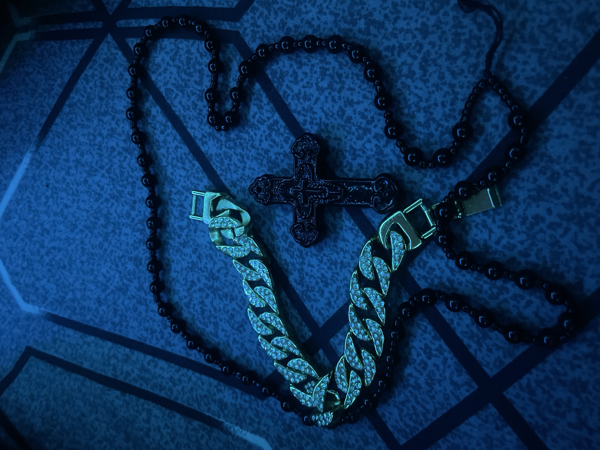 The image displays a dark, textured surface illuminated by blue light. Two necklaces are laid out on the surface. One is a rosary with black beads. The other is a gold-colored Cuban link chain adorned with rhinestones. A dark, ornate cross lies between the two necklaces, positioned to the right of the chain and near the rosary. The lighting casts shadows from the objects onto the surface, creating a dramatic effect.