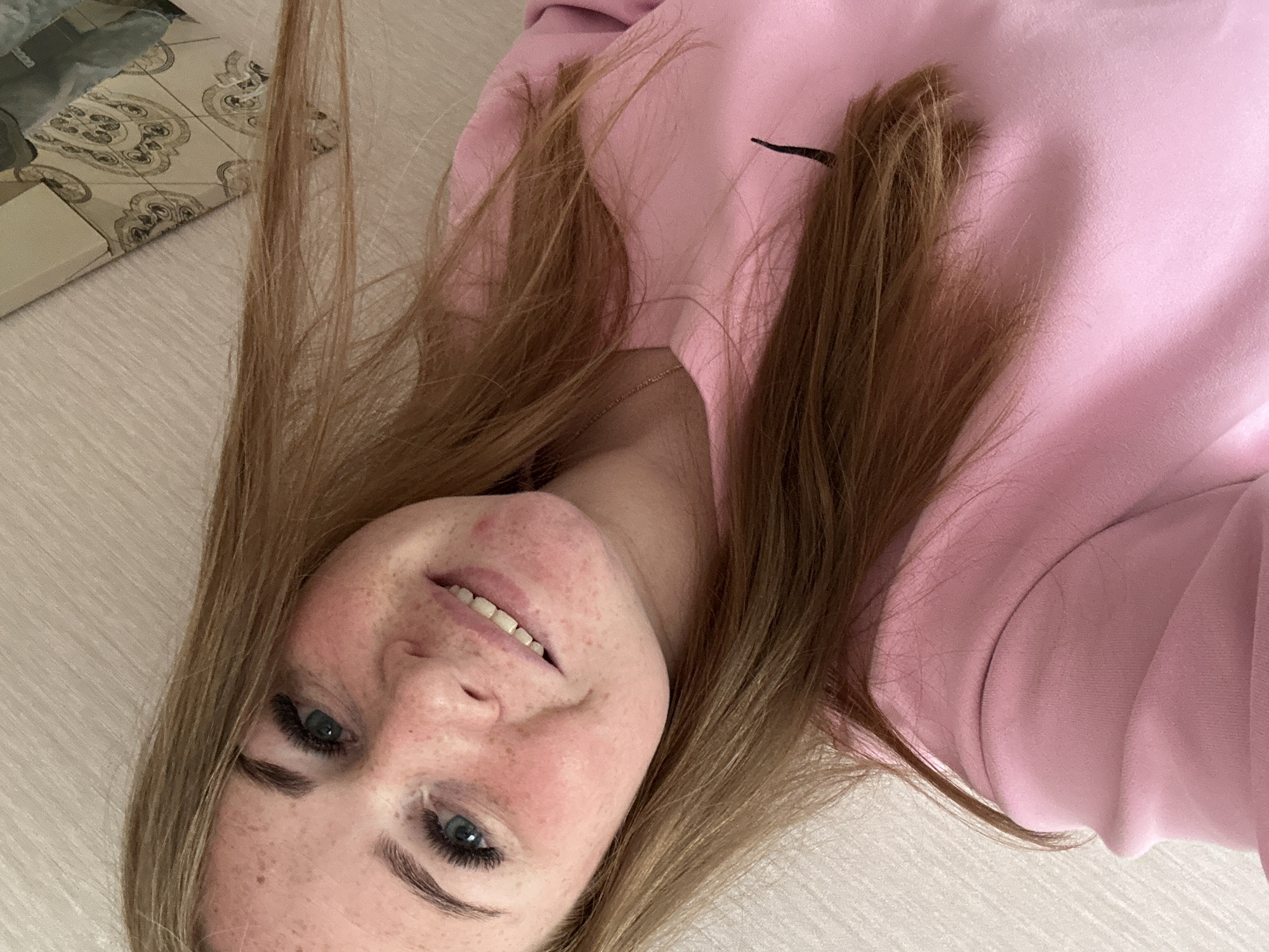 A young woman with fair skin and light brown hair lies sideways on a light-colored, possibly wooden, surface. Her face is tilted towards the camera, revealing blue eyes, freckles, and a slight smile. She wears a light pink sweatshirt with a black swoosh logo visible near the neckline. A portion of patterned tile is visible in the upper left corner of the image, possibly indicating the presence of furniture or objects in the background. The location is Chaikovsky, Russia.