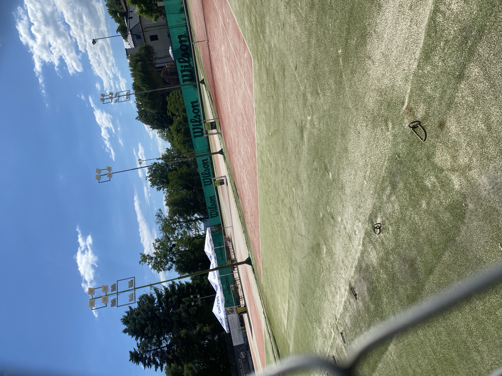 The image shows a tennis court with a green surface in the foreground and a red surface in the background. There are tennis nets and lampposts on the court. In the background, there are trees and buildings. The sky is blue with some clouds. The scene is outdoors on a sunny day. There are no people in the image.