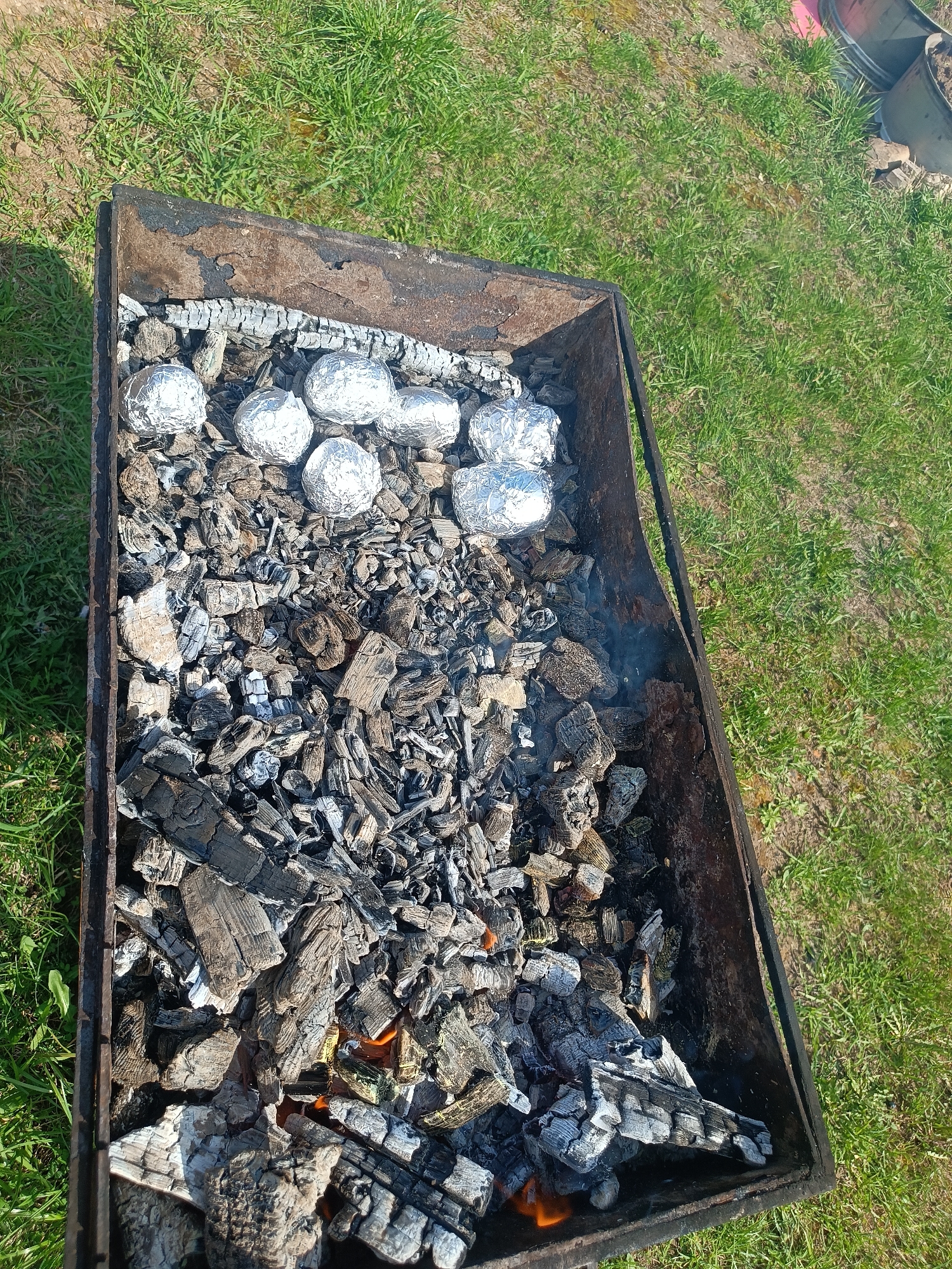 A top-down view shows a rectangular metal grill filled with charcoal and wrapped food on a grassy area. The grill is filled with wood charcoal that is partially burnt, with some embers still glowing. Six objects are wrapped in aluminum foil and placed along one edge of the grill. The grill sits on a lawn with short green grass. The image does not provide information to verify the location in Motyakovo, Russia. No people or interactions are evident in the image.