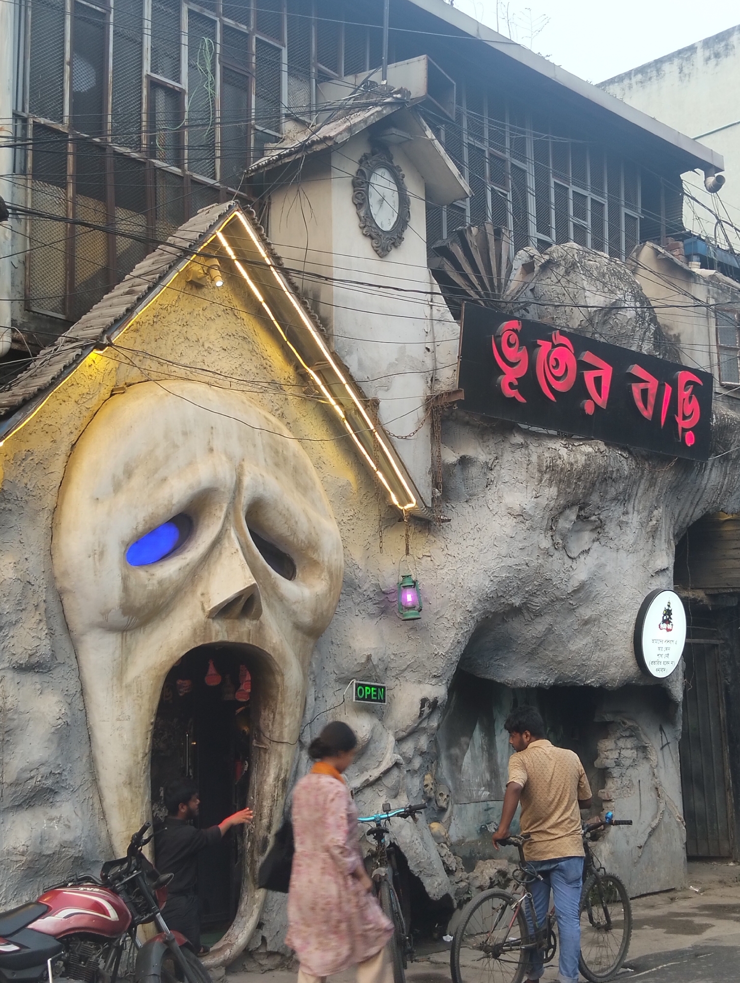 This image depicts the exterior of "Bhuter Bari," a horror-themed restaurant located in Dhaka, Bangladesh. The building is partially constructed with a large, sculpted face with a wide-open mouth serving as the entrance, and one eye is illuminated with a blue light. Another building is next to it with a sign in Bengali script, also sculpted to appear rocky, bearing the name of the restaurant. A small clock tower is affixed to the top of the building.
In front of the entrance, a man dressed in black stands at the doorway. A woman in a pink dress walks past the entrance. A motorcycle is parked near the entrance. To the right, a man wearing a yellow shirt and jeans stands with his bicycle. Another bicycle is nearby. The background shows a multistory building with barred windows, and an "OPEN" sign is visible near the restaurant's entrance. Electrical wires are overhead.