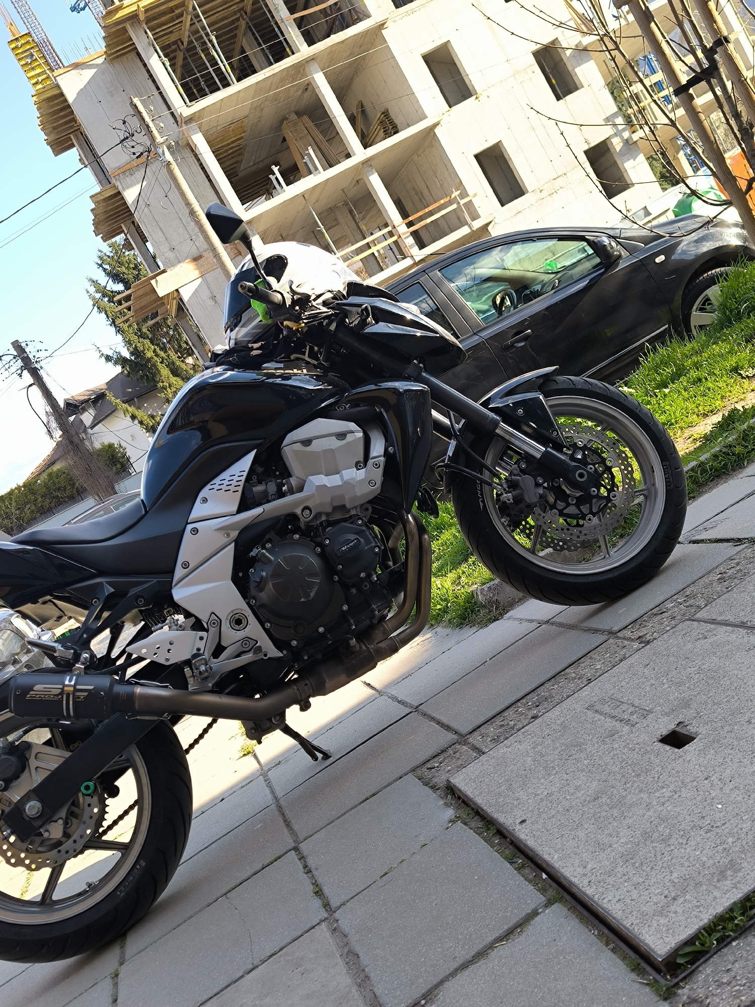 The image shows a black Kawasaki Z750 motorcycle parked on a paved sidewalk. A white helmet sits on the motorcycle. A black car is parked in the background on a patch of grass. Behind the car is a building under construction. The location appears to be an urban setting in Sofia, Bulgaria, judging by the architecture and paving. There is no visible human interaction in the image. The day is sunny, casting shadows on the pavement.
