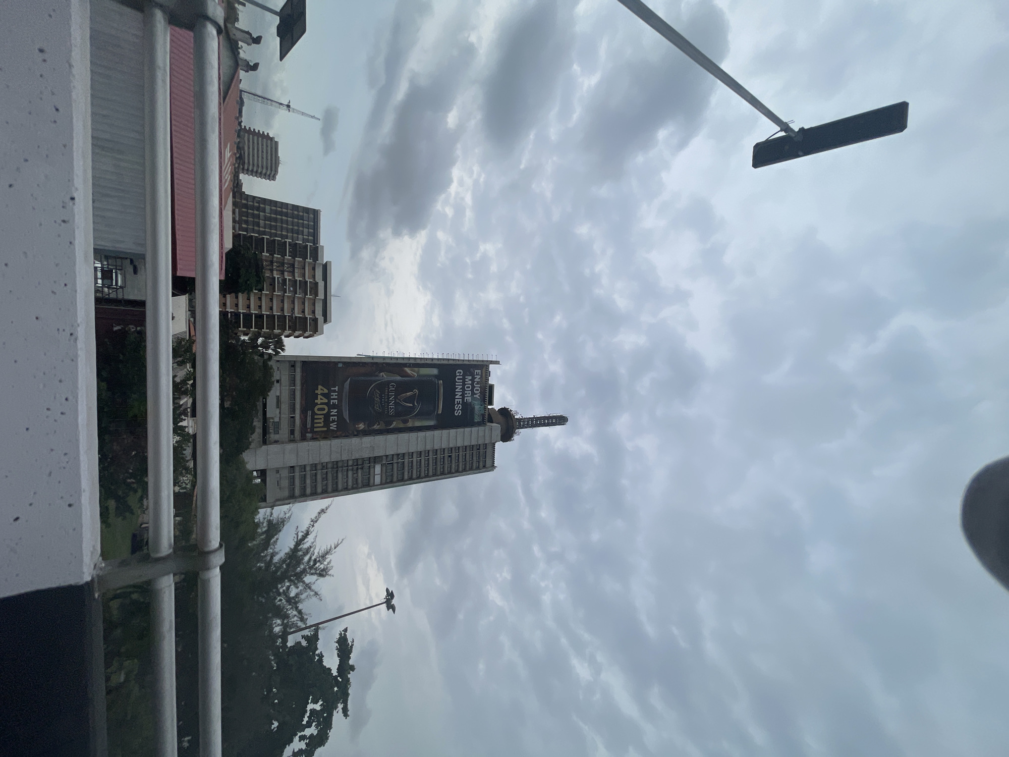From a low vantage point, the image shows a cloudy sky over Lagos, Nigeria. Several buildings are visible, including a tall skyscraper adorned with a large advertisement for Guinness beer. In the foreground, there is a metal railing with a section of a concrete structure. A lamppost and overhead street lighting can also be observed. The overcast conditions create a muted and slightly somber atmosphere.