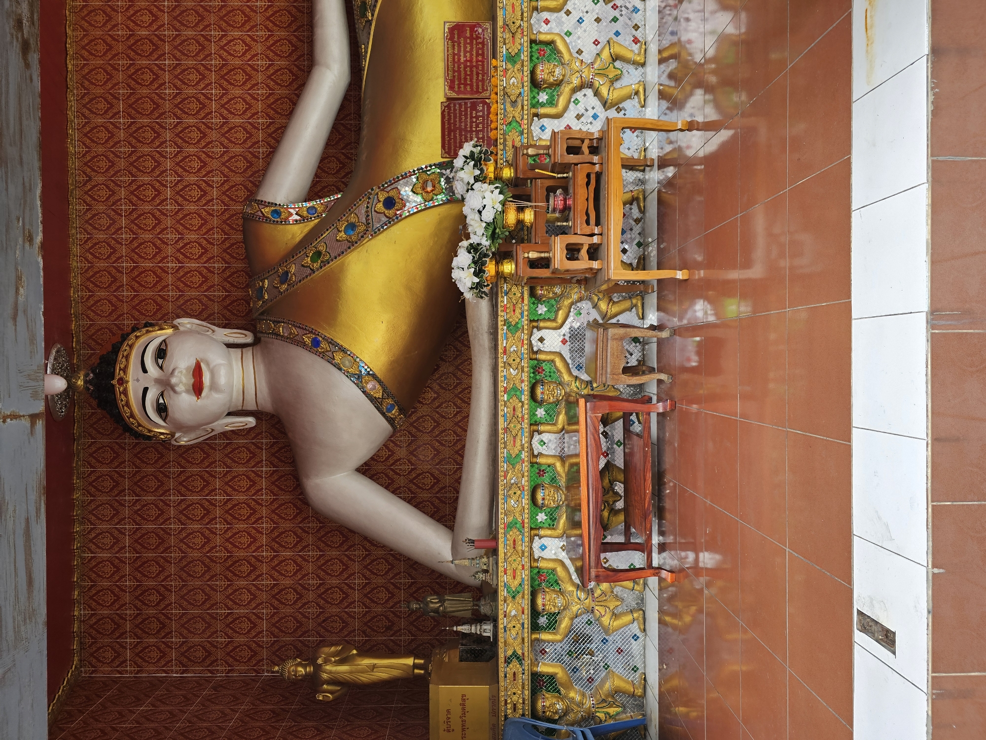 The image shows a large, reclining Buddha statue inside a temple in Bangkok, Thailand. The statue is white with golden robes and is lying on its side, resting its head on its arm. The background wall is covered in a red, patterned wallpaper. In front of the statue, there is a low wall decorated with figures of men in golden attire holding up the platform the Buddha rests on. On this platform are several small wooden tables with offerings, including white flowers in vases. The floor is tiled with reddish-brown tiles that reflect the light. The scene appears to be an indoor space within a temple complex.