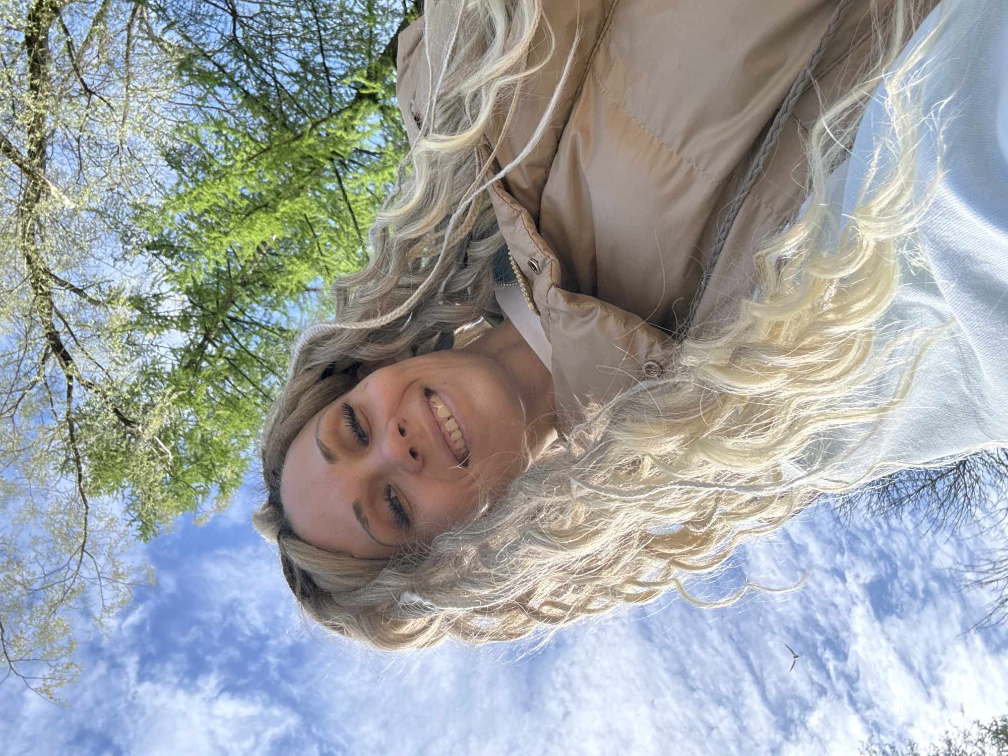 A woman is smiling at the camera with blue sky and trees in the background. The woman has blond, wavy hair. She is wearing a tan puffer jacket and a light blue sweater. The sky is blue with scattered clouds. There are trees with green leaves in the background. The photograph was taken outdoors during the day. Location cannot be confirmed with provided information.