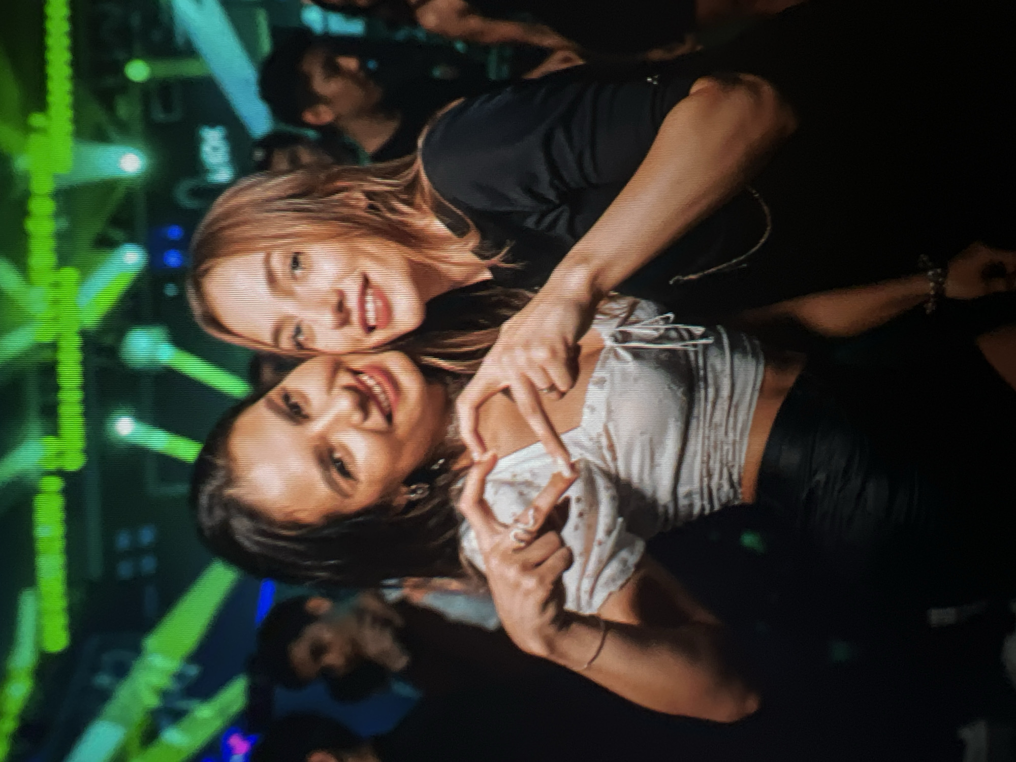 Two young women are posing for a photo in a dimly lit environment with green and blue lights in the background. The woman on the left has dark hair and is wearing a white top with a sweetheart neckline and a black skirt. She is making a heart shape with her hands. The woman on the right has blonde hair and is wearing a black top. She is smiling and her arms are around the other woman's shoulders. Several other people are visible in the background, blurred and out of focus. The scene appears to be a nightclub or a party. The location of the Qarağandı city administration in Kazakhstan is not discernible from the image.