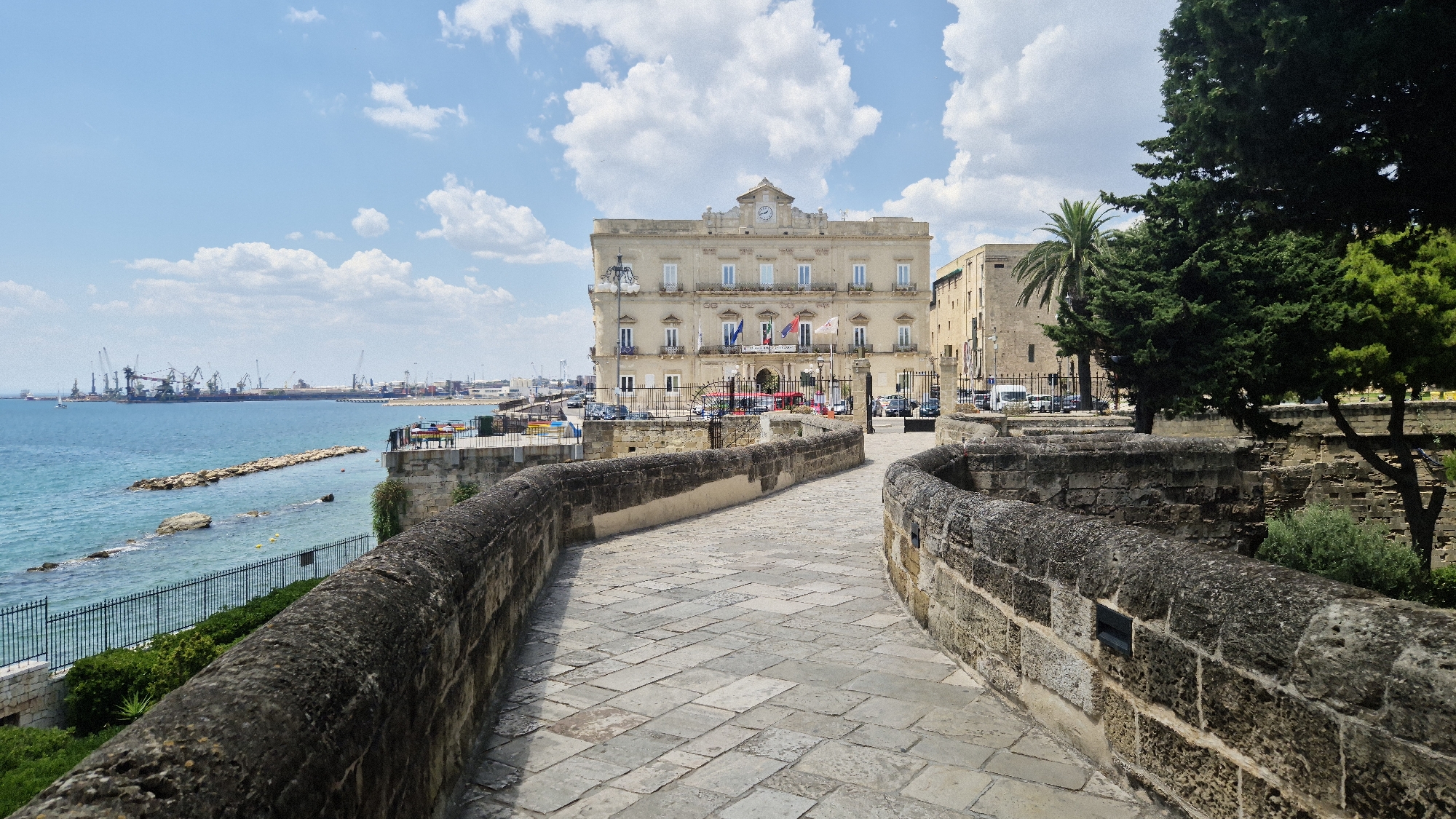 This is a landscape photograph of the Lungomare Vittorio Emanuele in Taranto, Italy, featuring a view of the historic Palazzo Amati and the port in the distance. A paved stone walkway, lined by weathered stone walls, leads towards the multi-story Palazzo Amati, a light-colored building with numerous windows and flags displayed on its facade. The building has a clock on its roof and is situated against a bright blue sky with scattered white clouds. To the left of the walkway, the blue waters of the Ionian Sea are visible, with a distant view of the Taranto port, characterized by numerous cranes and industrial structures. The foreground is dominated by the stone walls and the walkway, with a large, dark green tree on the right side of the frame. No people are clearly visible in the frame. The lighting suggests it is daytime, possibly late morning or early afternoon.