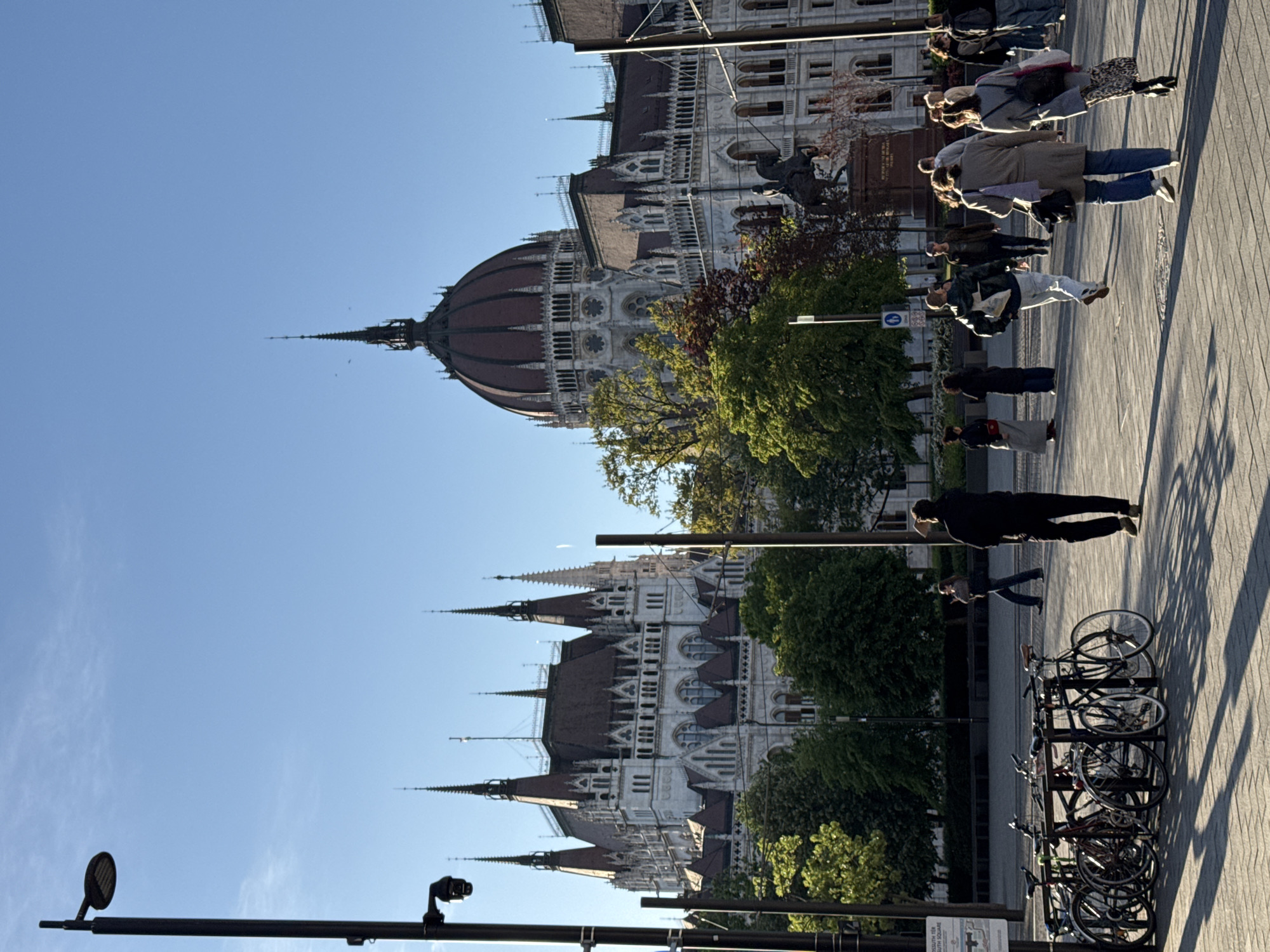 This image captures a vibrant street scene in Budapest, Hungary, featuring the iconic Hungarian Parliament Building. The grand gothic revival structure, with its distinctive central dome, numerous spires, and intricate white stone facade, dominates the left side of the frame under a clear blue sky. Parts of its dark red roof are visible. Green trees, some showing early signs of autumn with reddish leaves, partially obscure the lower sections of the building.
On the right, a wide, paved public square or promenade is populated by several pedestrians. Individuals and small groups are seen walking, and their long shadows stretch across the ground, suggesting the golden hour of either early morning or late afternoon. Their clothing is casual. In the foreground on the right, a bicycle rack holds multiple parked bicycles. An equestrian statue is visible on the plaza closer to the Parliament.
Various black street poles, some equipped with surveillance cameras or lighting, are scattered throughout the image. A sign on one pole in the lower right corner is partially legible, displaying "KOSSUTH TÉR" and an "M" symbol, confirming the location as Kossuth Square and indicating a nearby metro station. The overall atmosphere is calm, depicting a typical day with people enjoying the public space around a famous landmark.