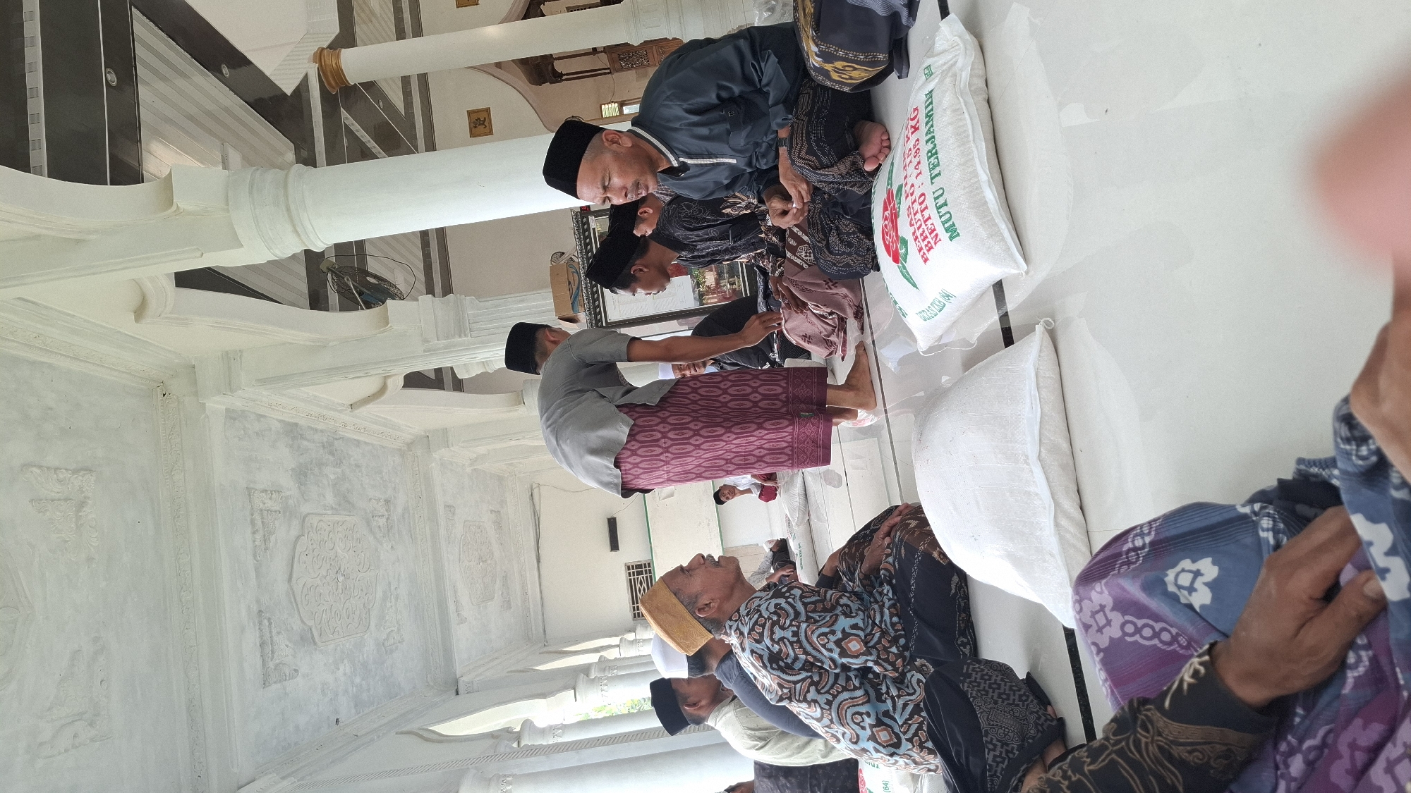 In a covered outdoor area, possibly a mosque or community hall, in Grong-grong, Indonesia, a group of men are gathered, seated on a white tiled floor. The men are dressed in traditional Islamic attire, including peci caps and sarongs. One man, wearing a grey shirt and a patterned maroon sarong, stands and appears to be interacting with the seated individuals. In the foreground, two large white sacks, presumably containing rice, are visible, with one sack displaying Indonesian text that reads "BERAS MUTU TERJAMIN". The architectural style features white columns and ornate white ceiling decorations, suggesting a well-maintained structure. The lighting appears to be natural, indicating daytime. The overall scene depicts a communal gathering, possibly for a religious event or distribution of aid.
