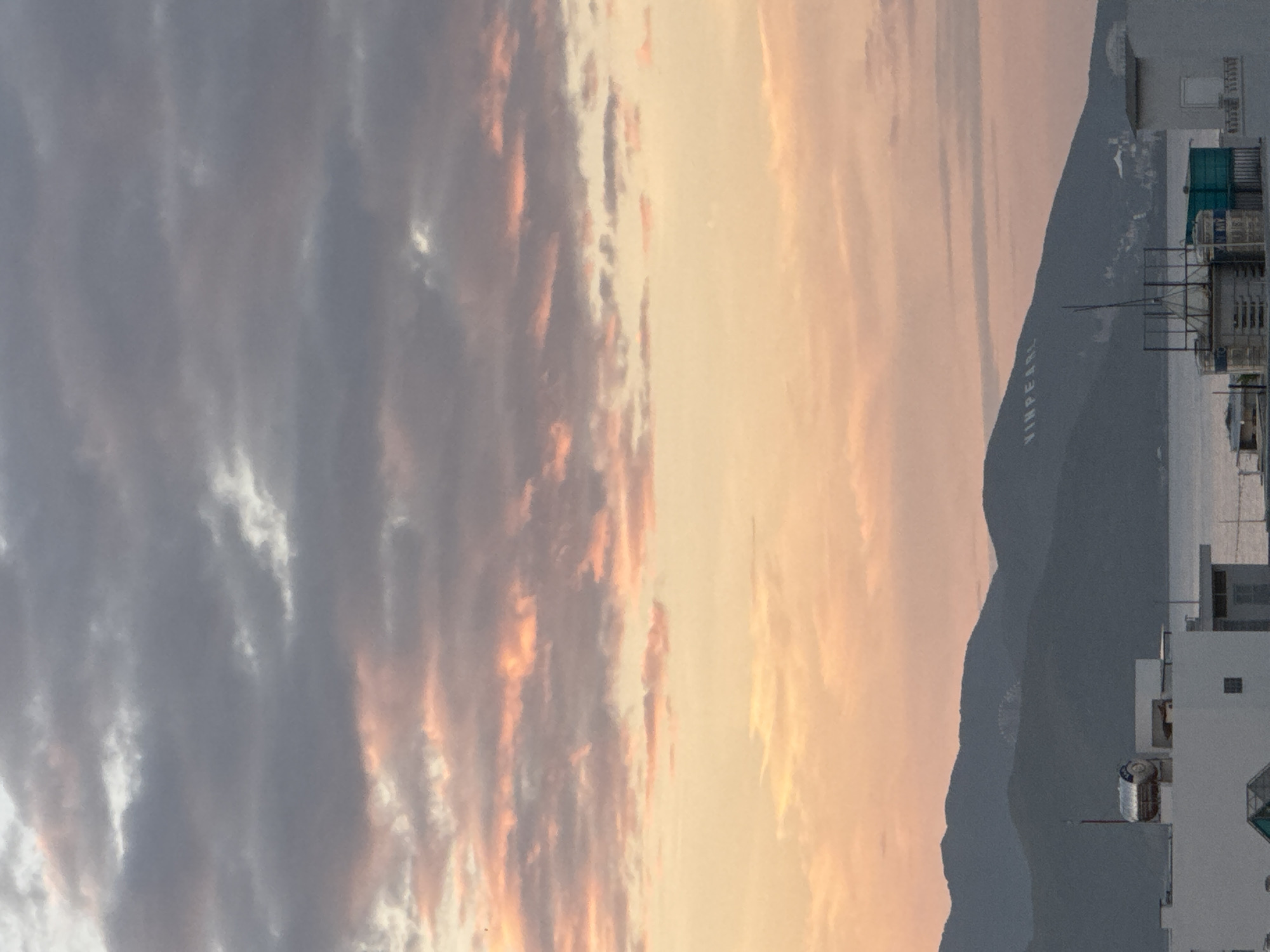 The image captures a coastal city during what appears to be sunset or sunrise, with a dramatic sky filled with hues of pink, orange, and gray clouds. In the foreground, the rooftops of buildings are visible, one with a prominent silver water tank. Beyond the buildings, a body of water, likely the sea, stretches out to meet a range of dark, silhouetted mountains. A notable detail on the mountainside is the large white lettering spelling "VINPEARL," indicating the location as Nha Trang, Vietnam, and likely referring to the Vinpearl resort complex situated on Hon Tre Island or a similar development. The overall scene depicts a tranquil, scenic view of a coastal urban landscape.