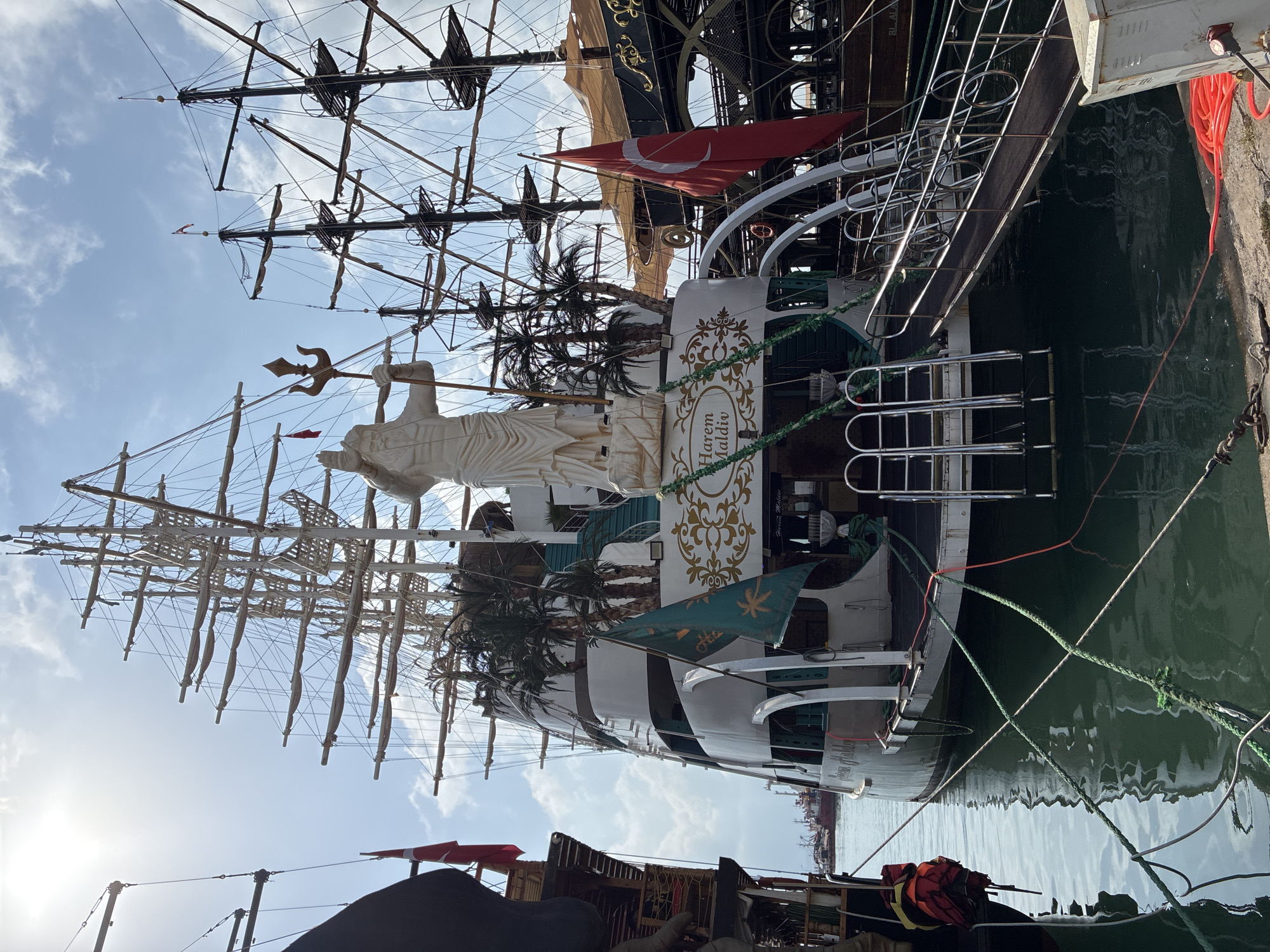 The image captures a view of a large sailing ship docked in the harbor of Alanya, Turkey. The ship features multiple masts and sails, indicative of a traditional sailing vessel. Prominently displayed on the stern of the ship is a large white statue of a bearded man, possibly a deity or historical figure, holding a trident. The statue is positioned above decorative elements and palm trees. The ship also displays flags, including a red Turkish flag with a white crescent and star. Various ropes are securing the ship to the dock, and there are gangways for access. In the background, other boats and structures can be seen, suggesting a busy port environment. The water is calm, reflecting the sky and the ships. The overall scene depicts a maritime setting in Alanya.