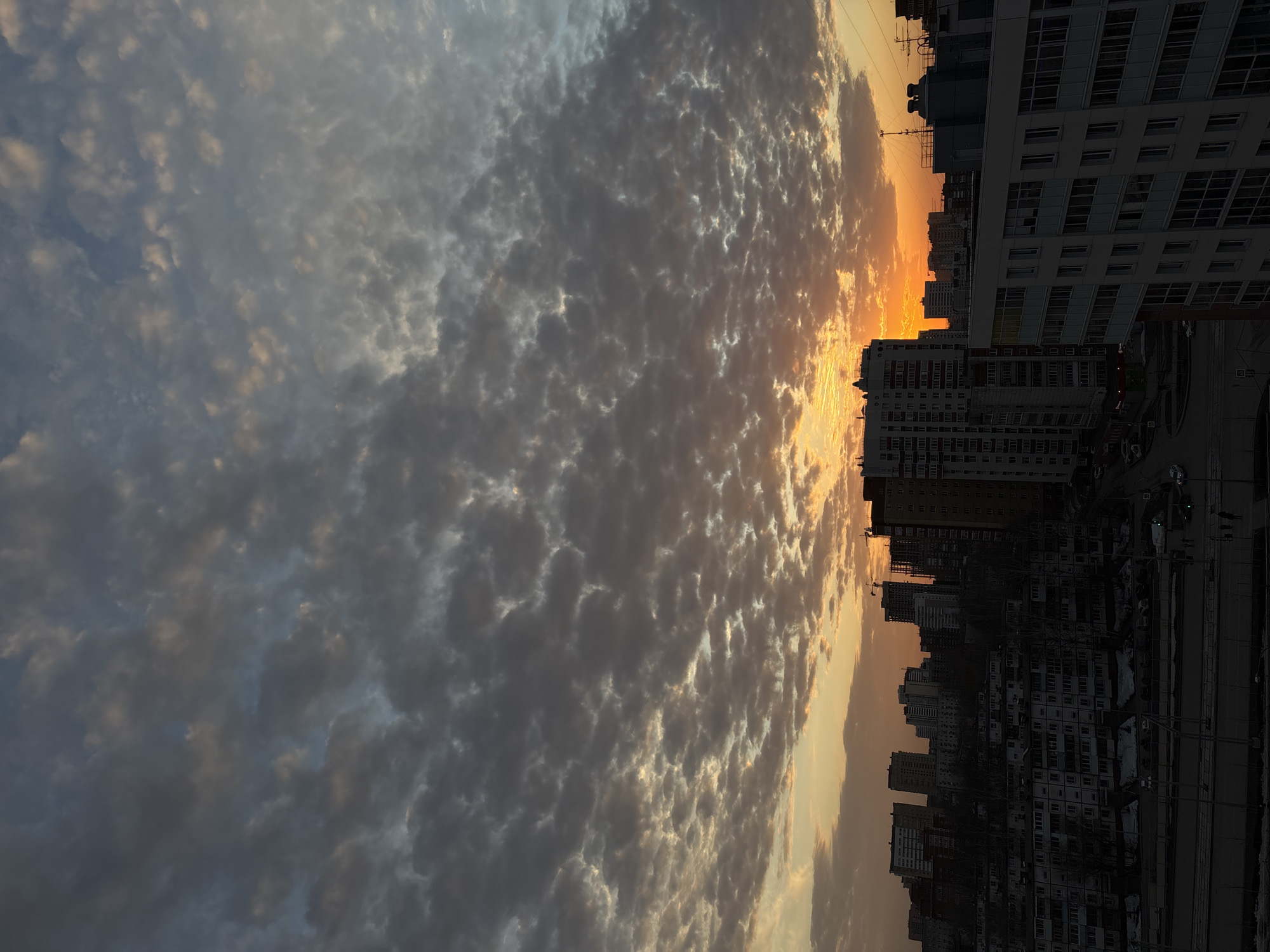 The image captures a sunset in Perm, Russia. The sky is filled with altocumulus clouds, colored with shades of gray, blue, and orange from the setting sun. The cityscape in the foreground is composed of multi-story residential buildings. A road and vehicles are visible along the street. Construction cranes are also visible, indicating ongoing development in the area. The time of year appears to be late winter or early spring due to the presence of some remaining snow.