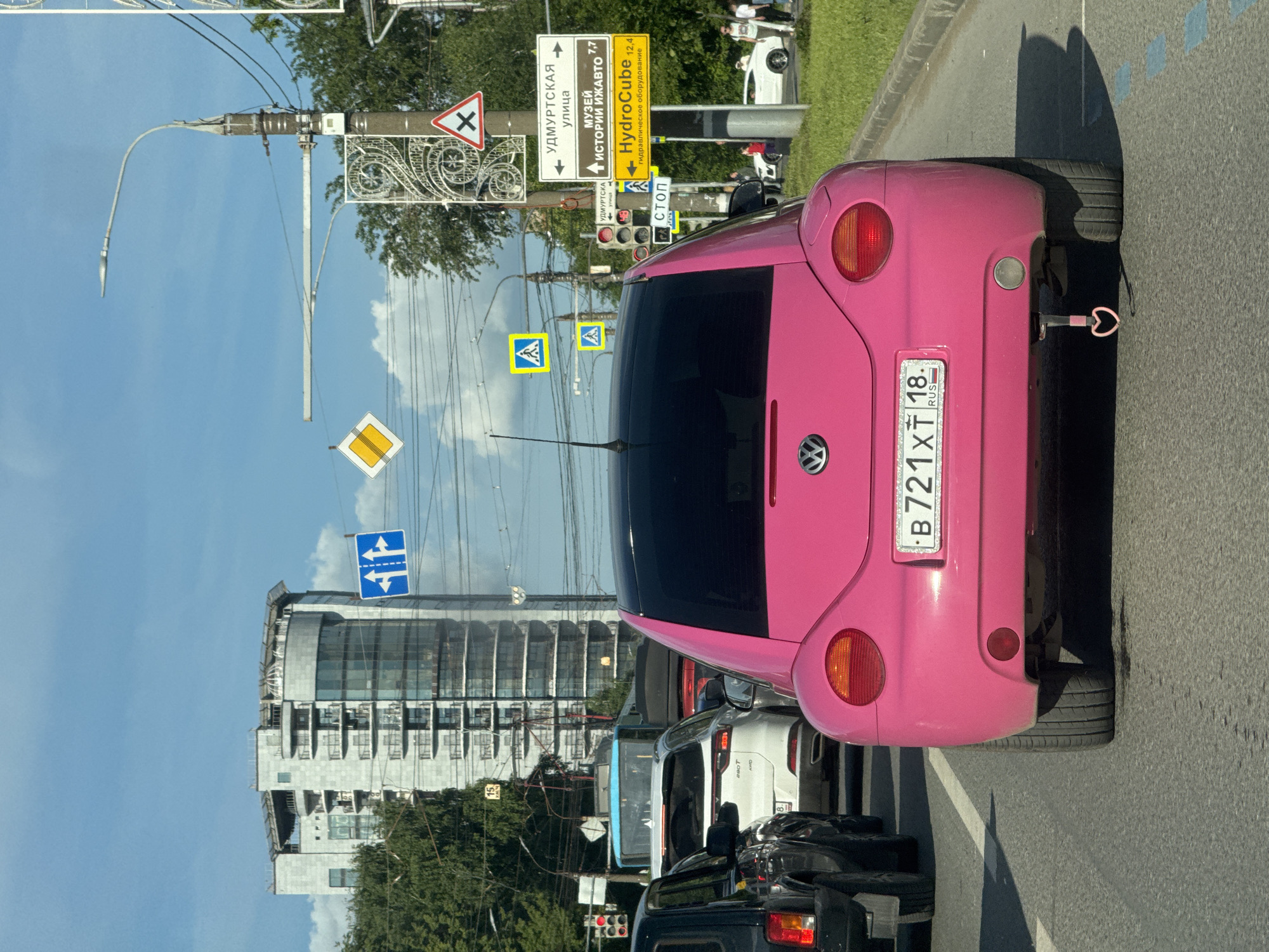 A vibrant pink Volkswagen Beetle is the central subject of this daytime street scene in Izhevsk, Russia. The car is seen from the rear, featuring a distinctive heart-shaped tow hitch and a Russian license plate "B721XT 18 RUS," which designates the Udmurt Republic. It appears to be stopped in traffic, with other vehicles visible ahead and to the left.
The setting is an urban street on a sunny day with a clear blue sky and some white clouds. To the left, a modern building with a glass facade and multiple balconies stands in the background, partially hidden by green trees. Overhead, a network of wires is supported by a street lamp pole.
Several Cyrillic road signs are prominent. A large yellow sign indicates "УДМУРТСКАЯ улица" (Udmurtskaya street) pointing left, "МУЗЕЙ ИСТОРИИ ИЖАВТО 7,7" (Izhavto History Museum 7.7 km) pointing straight, and "HydroCube 12,4" (HydroCube 12.4 km) for "гидравлическое оборудование" (hydraulic equipment) pointing right. Other signs include a small "СТОП" (STOP) sign, a yellow diamond indicating a priority road, a red triangular crossroads warning sign, and blue and yellow signs related to pedestrian crossings or road directions. A traffic light showing red is also visible, consistent with the vehicle being stationary.