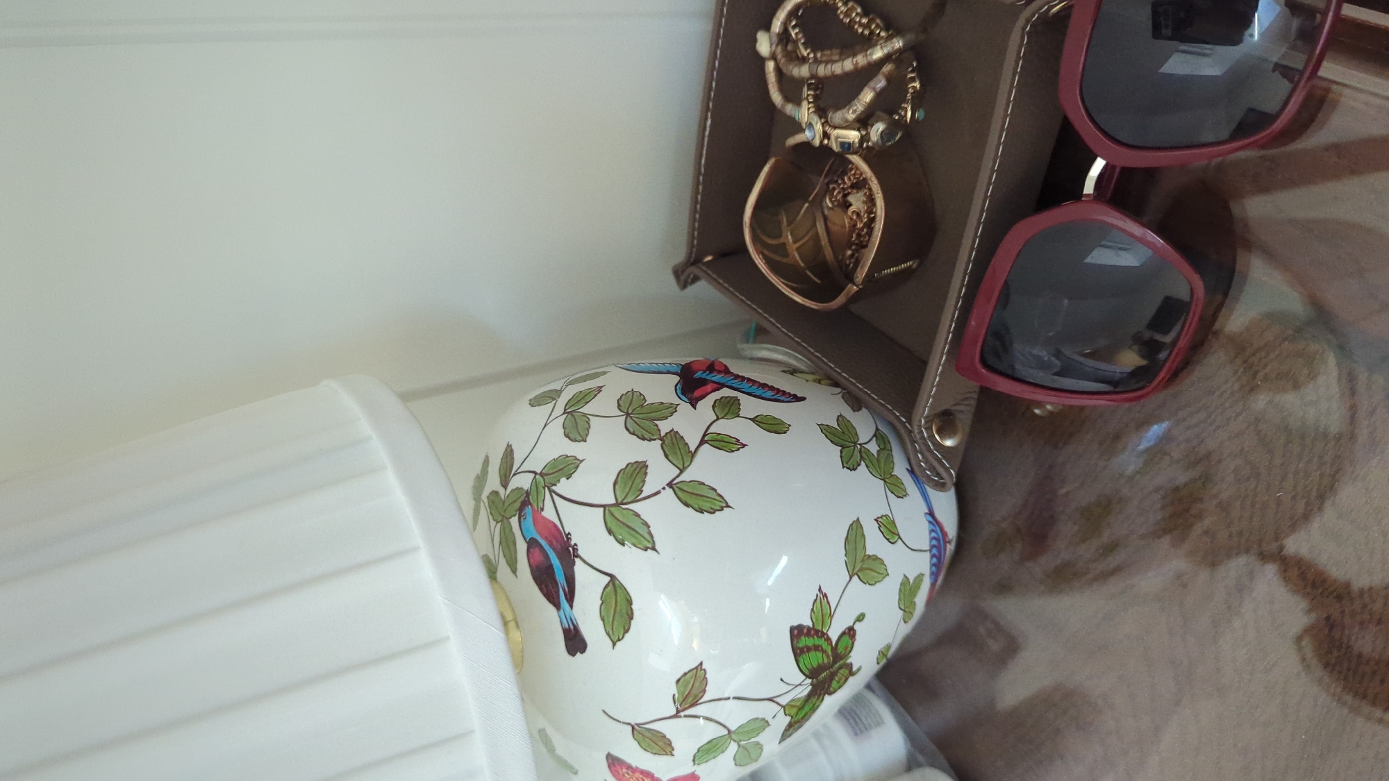 The image shows a close-up of a table surface. On the table, there is a white ceramic vase decorated with birds and foliage. To the right of the vase, there is a brown leather tray containing jewelry, including necklaces and bracelets. In the foreground, a pair of dark red sunglasses with large lenses rests on the table. The background is a plain white wall with vertical paneling. There is no indication of the city of Larkollen, Norway in the image.