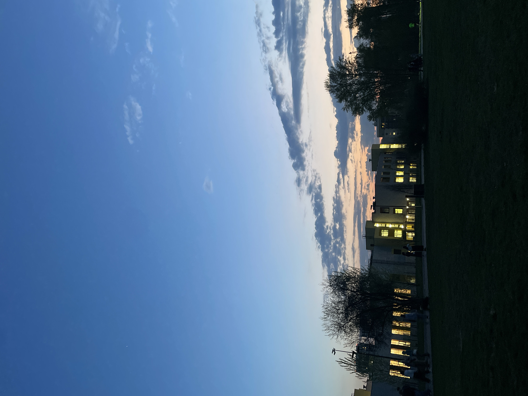 The image shows an evening scene in Kazan, Russia, featuring a park area with illuminated buildings along the edge of a grassy field. In the sky, long streaks of clouds stretch vertically, with the light reflecting the setting sun. Buildings with multiple lit windows suggest offices or apartments. Trees are silhouetted against the colorful sky. People can be seen near the buildings, but they are indistinct. The foreground consists of a darker green grassy area, likely a park.