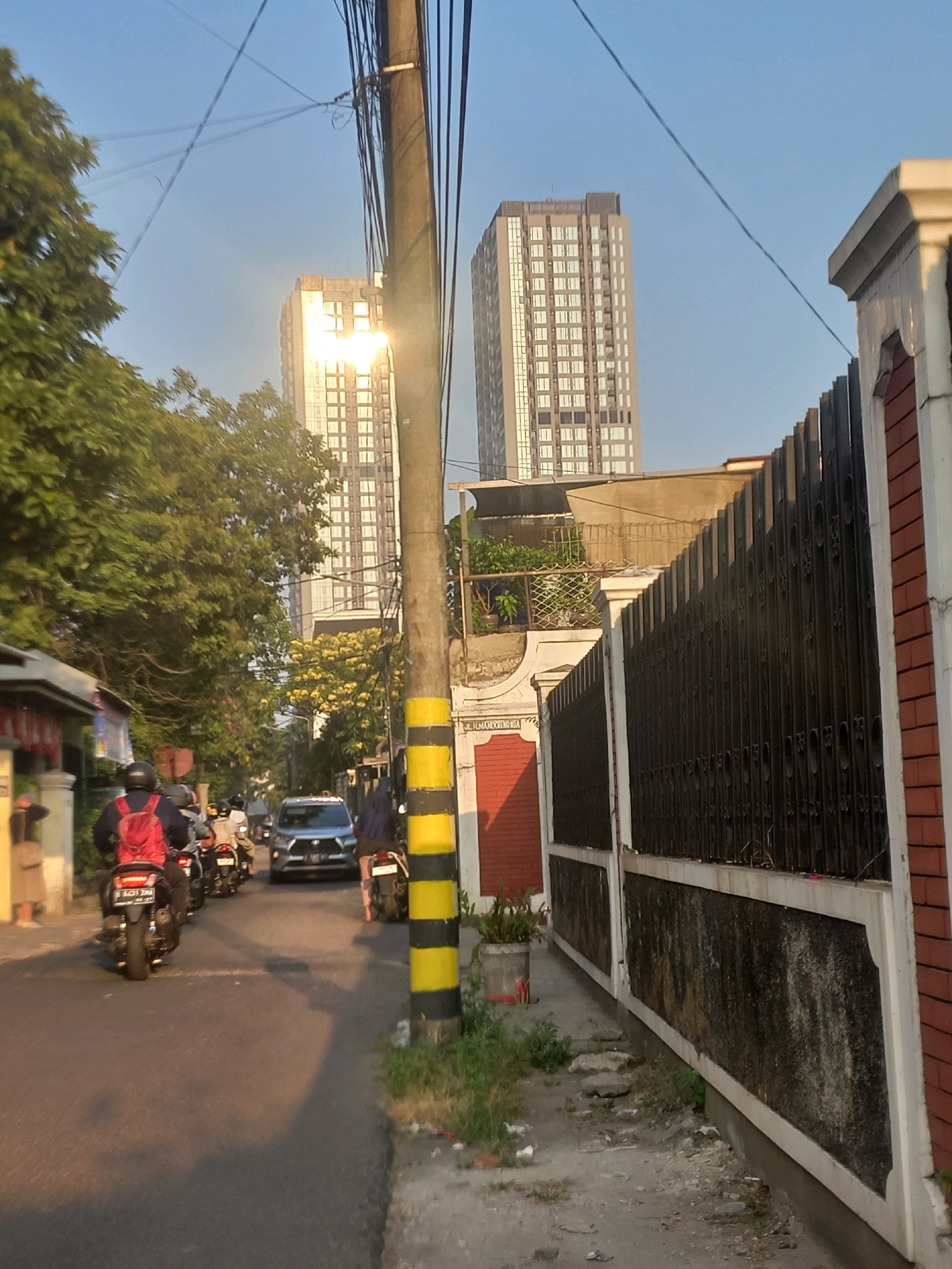 The image depicts a street scene in South Jakarta, Indonesia, during what appears to be late afternoon or early morning due to the lighting. In the foreground, a paved road is visible, with several motorcycles traveling on it. One motorcycle in the immediate foreground has a rider wearing a red backpack. Further down the road, a silver car is in motion. On the right side of the street, a tall, dark metal fence runs along the property line, bordered by a brick wall. A utility pole with black and yellow stripes stands prominently in the middle ground. Behind the fence and houses, two modern, multi-story buildings dominate the skyline, their glass facades reflecting the sunlight. Lush green trees and some flowering bushes are present, particularly on the left side of the road, adding a natural element to the urban environment. The sky is clear and blue.