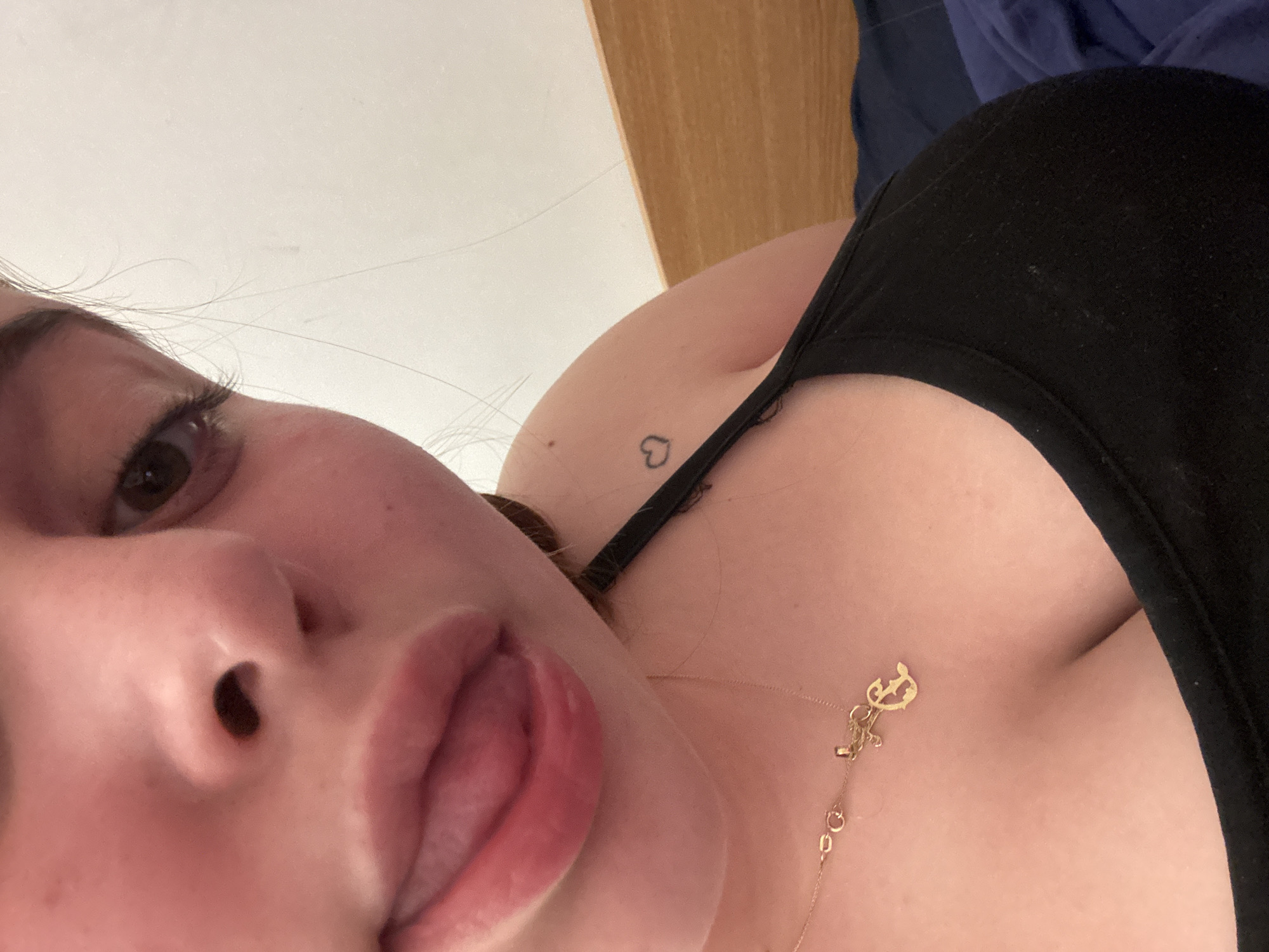 A close-up selfie shows a fair-skinned person wearing a black tank top. The person's face is the primary focus, with one eye visible, large pink lips slightly parted and tongue showing. A thin gold chain necklace with a decorative pendant lies on the person's chest. A small heart tattoo is visible on the upper chest near the shoulder. The background includes part of a wooden structure and a blue fabric. The location of Troyan, Bulgaria is not discernible from the image.