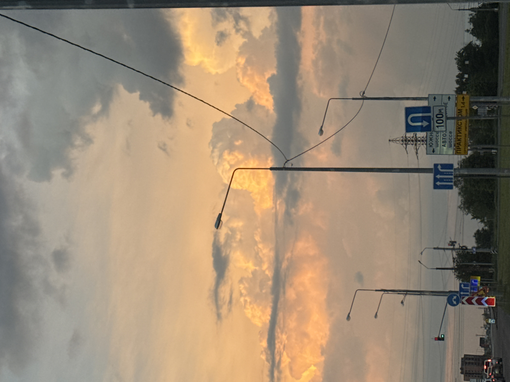 This image captures a sunset over a city intersection. The sky is filled with dramatic clouds illuminated by the setting sun, casting an orange glow on the scene. Several streetlights are visible, some silhouetted against the colorful sky. A traffic light is showing red, indicating that vehicles should stop. Road signs are present, including directional signs and a sign indicating "100m". Cars are visible on the road, suggesting ongoing traffic. Power lines and a transmission tower are also present in the background, indicating urban infrastructure. The overall scene depicts a typical city environment during twilight, characterized by a blend of natural light and man-made elements.