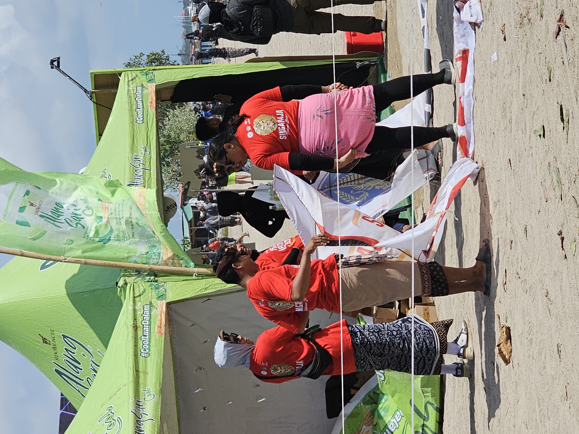 The image depicts an outdoor event, likely a festival or competition, set on a sandy ground near a body of water, possibly a beach or harbor. In the foreground, three people are visible. Two men, wearing red t-shirts and traditional patterned sarongs, are examining a large, partially unfurled kite. The kite is predominantly white with red and blue accents. To the right, a woman, also in a red t-shirt and a pink sarong with black leggings, stands facing away from the camera, looking towards the kite. Behind them, several green tents are set up, one prominently displaying the logo "Alang Sari Cool" and the hashtag "#CoolLuarDalam". In the background, a crowd of people are gathered, and some boats are visible in the water. The sky is partly cloudy. Based on the prominent "Alang Sari Cool" branding and the general atmosphere, this event could be taking place at Padang Galak Beach in Denpasar, Bali, Indonesia, which is known for kite festivals.