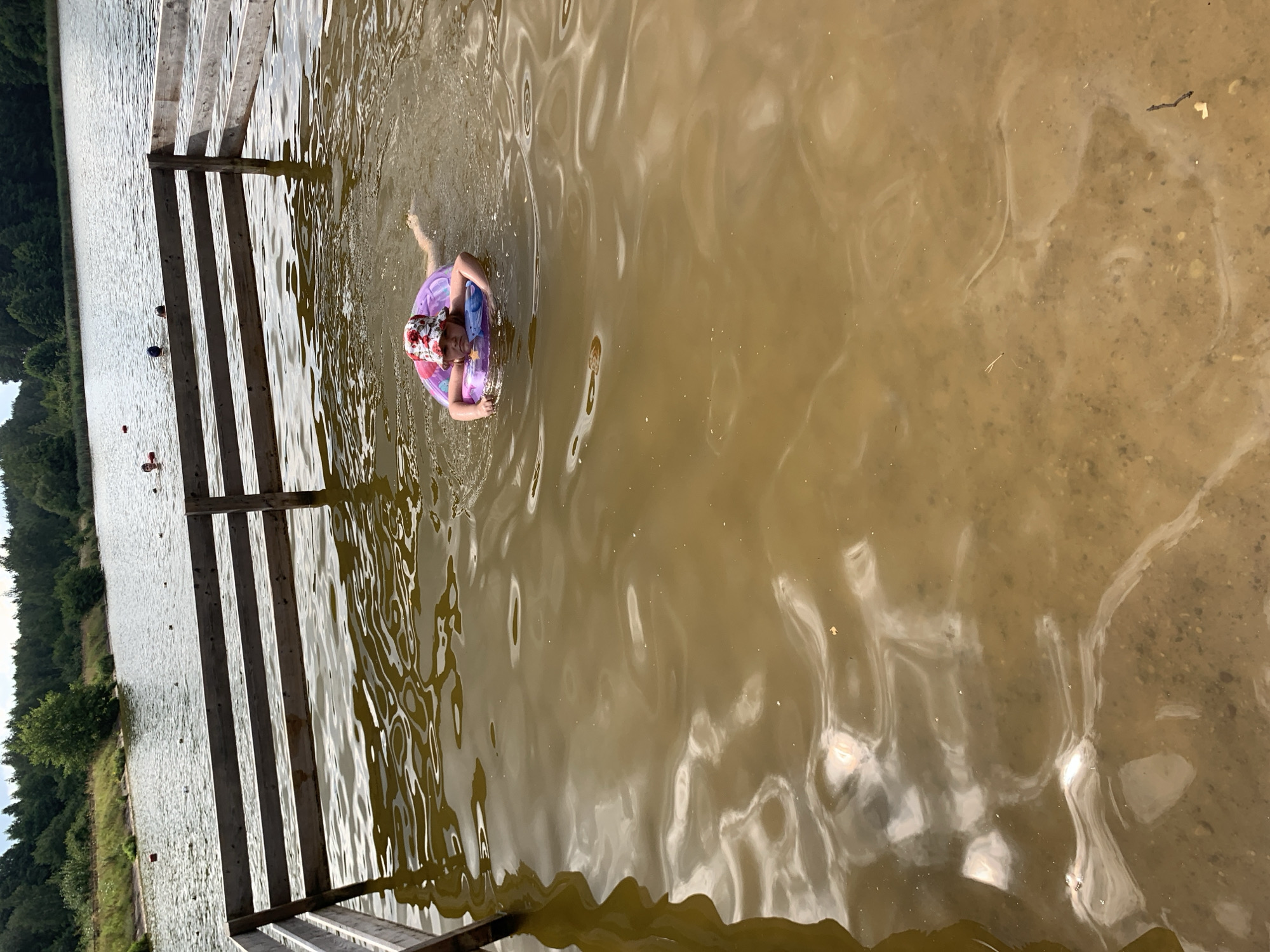 A young child wearing a floral hat swims in a lake, holding onto a pink inflatable ring. The water is murky and brown, with reflections of the sky and trees on its surface. A wooden fence runs along the edge of the water, separating it from a grassy bank lined with trees in the background. The sky appears overcast. The location is identified as Грозаўскі сельскі Савет, Belarus.